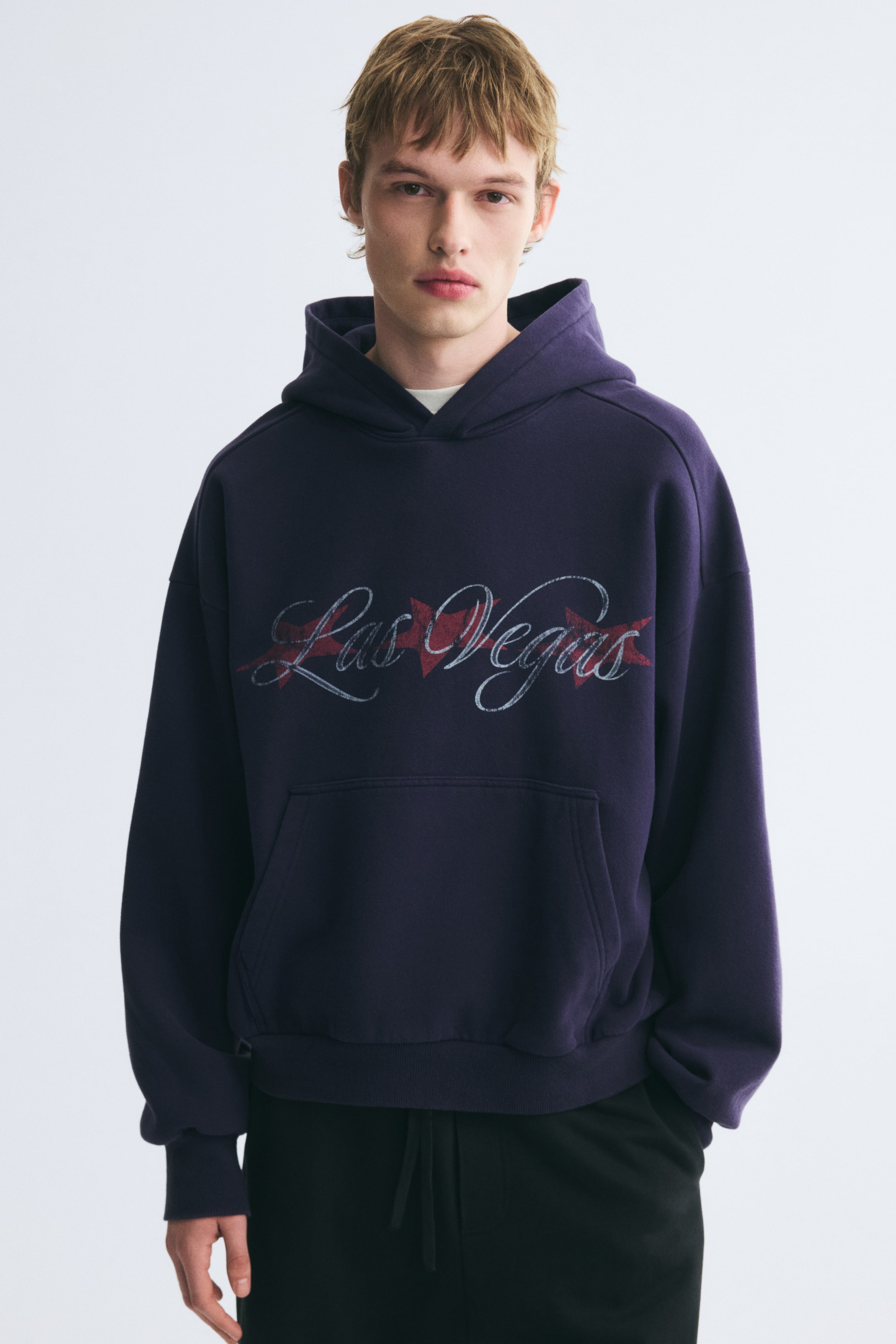 View larger image: An oversized, dark purple hoodie featuring a graphic print of "Las Vegas" in cursive script across the chest is being worn by the model. The hoodie features dropped shoulders, a kangaroo pocket, and ribbed cuffs, and is paired with black drawstring pants.
