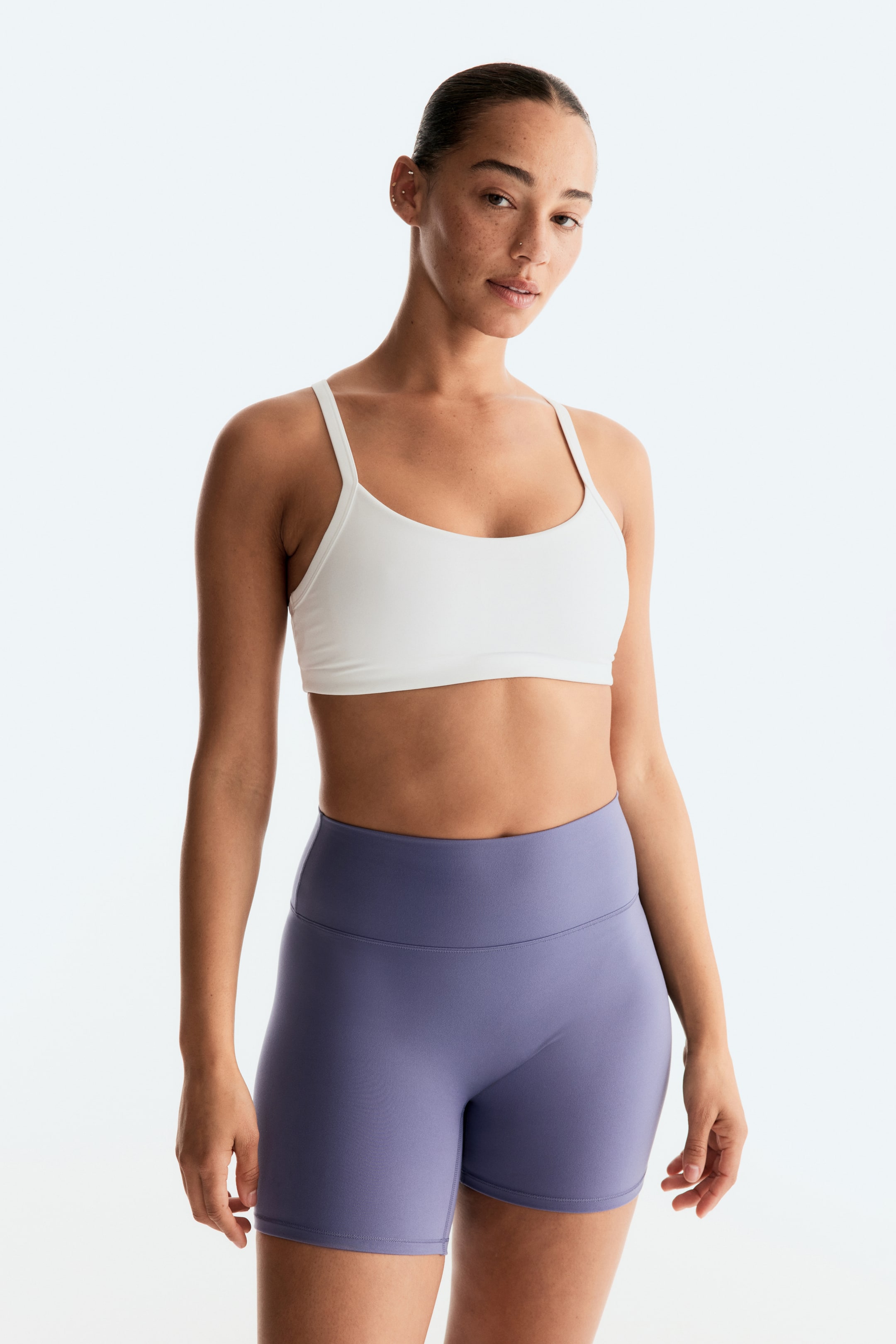 Ver imagen más grande: Un sujetador deportivo blanco con escote redondo y tirantes finos se lleva con unos pantalones cortos atléticos morados de tiro alto y longitud hasta el muslo, ambas prendas ajustadas al cuerpo.