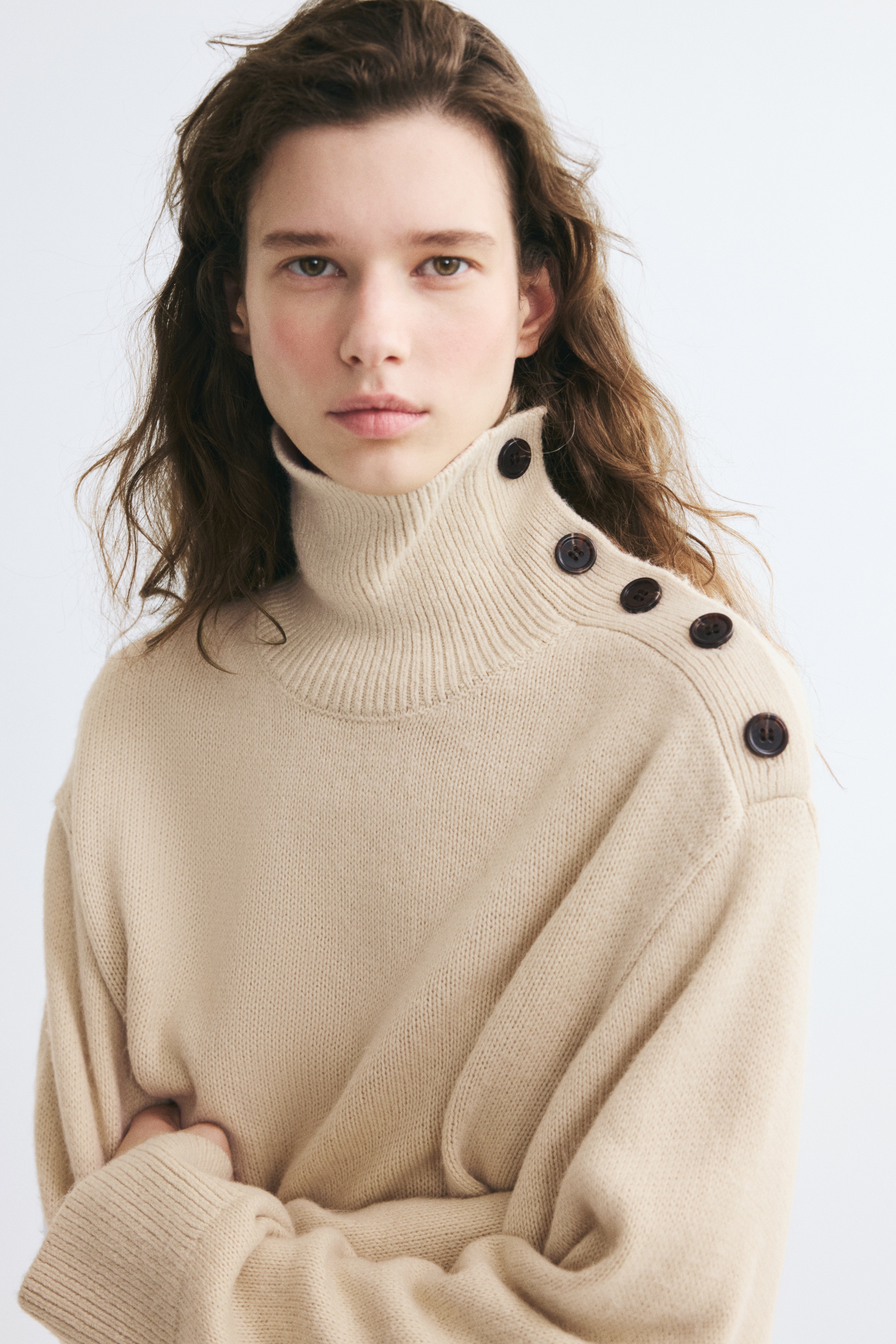 A cream-coloured knit sweater is shown from the front, featuring a high ribbed collar with a diagonal button placket along the left shoulder. The sweater features dropped shoulders, long sleeves with wide ribbed cuffs, and a matching ribbed hem.