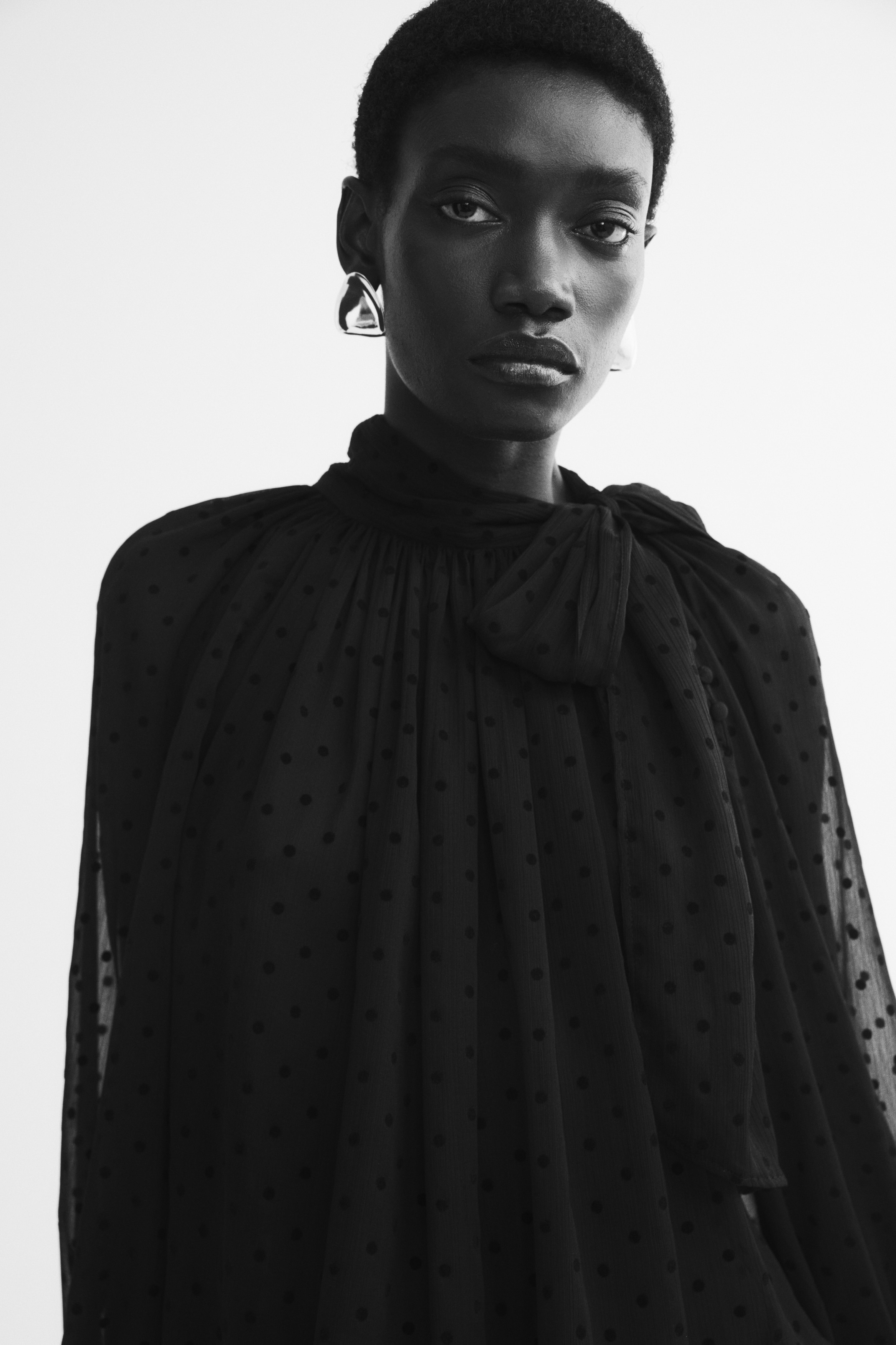 Una blusa negra transparente de lunares está vista de frente, con un cuello alto fruncido y un gran lazo lateral para anudar. La blusa presenta una silueta holgada y plisada, con mangas largas abullonadas que se estrechan hasta unos puños anchos y estructurados.
