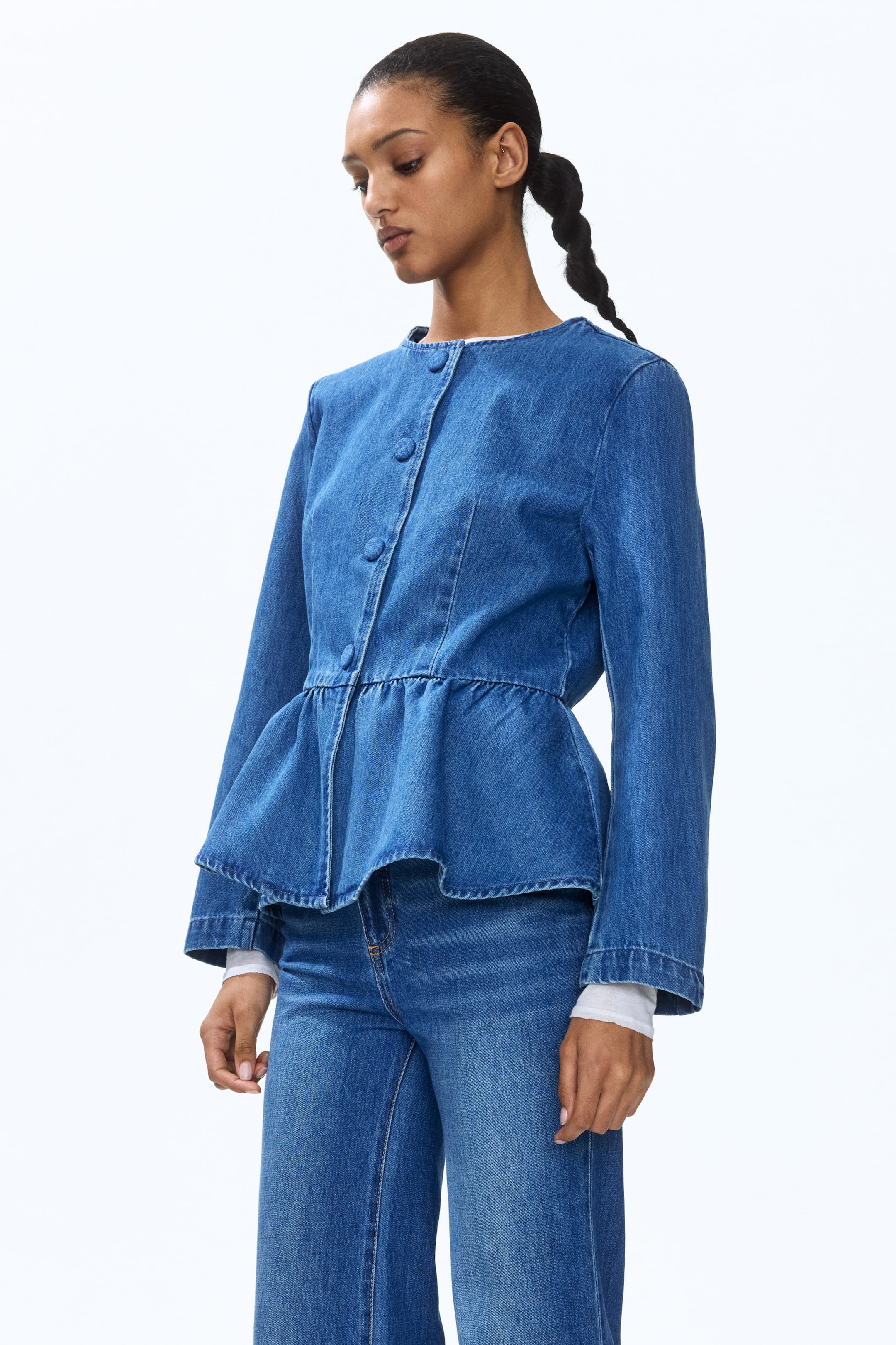 View larger image: A blue denim peplum jacket is being presented, angled slightly to the right, showcasing a band collar, long sleeves, a front button closure with circular buttons, and a gathered peplum hem. A white long-sleeve top is visible underneath, and the jacket is paired with matching denim wide-leg trousers.