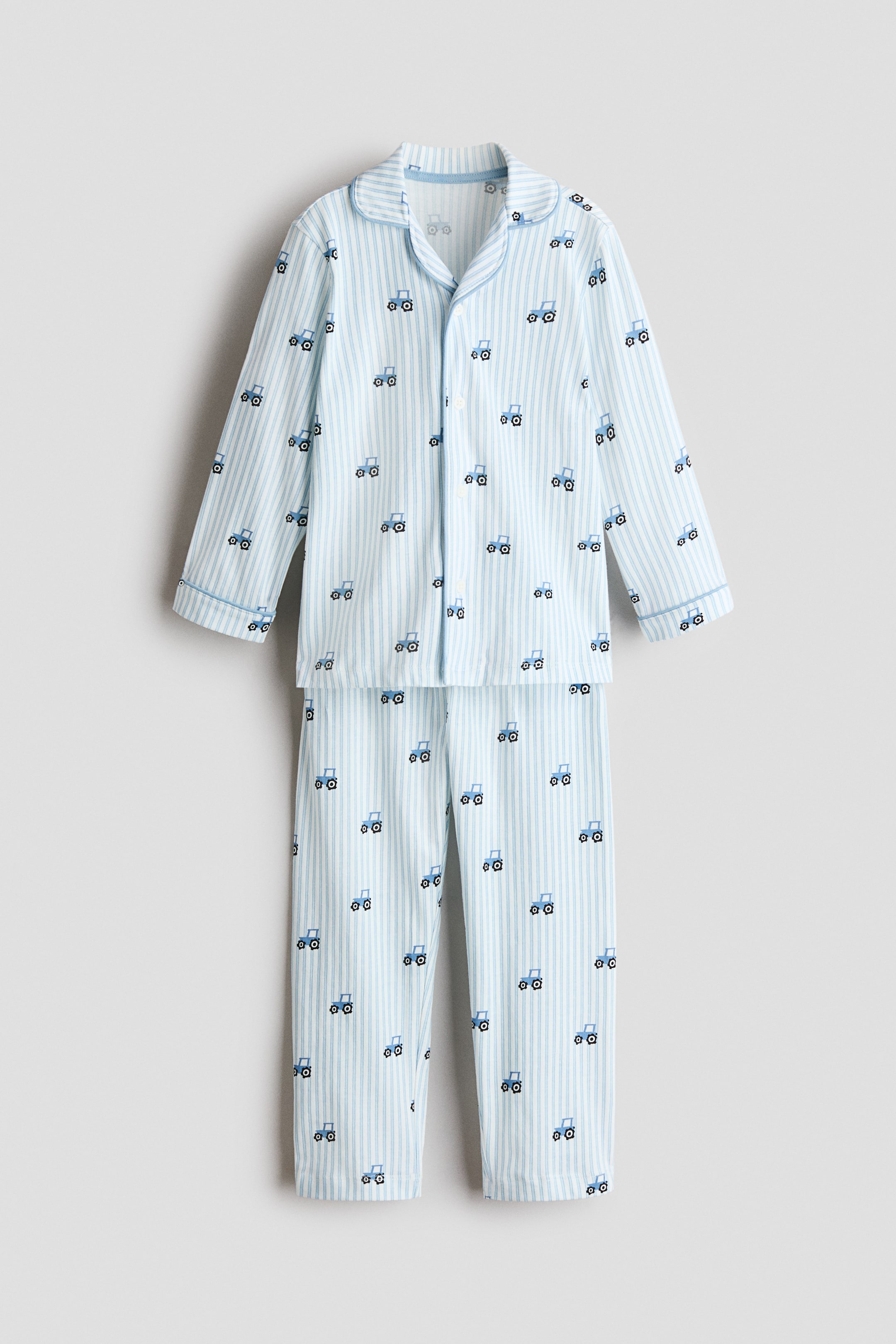 View larger image: A two-piece pajama set features a light blue and white vertical stripe pattern, accented with repeating small blue tractor motifs. The long-sleeved top has a full-button front and notch lapels, with light blue piping detail on the collar and cuffs, while the matching long pants complete the set.