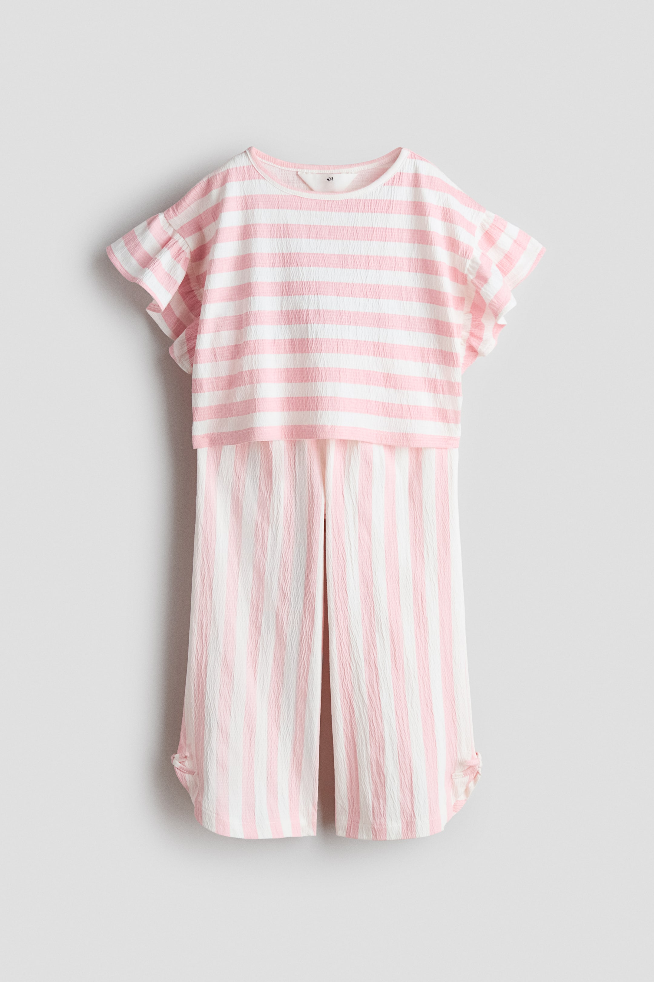 Visualizza immagine più grande: Un set composto da un top e pantaloni in tessuto jersey a righe rosa e bianche con texture, presentato frontalmente. Il top mostra righe orizzontali, un girocollo, spalle scese e maniche svolazzanti. I pantaloni presentano righe verticali, una vita elasticizzata con bordo arricciato e un piccolo spacco sul lato di ciascun orlo con bordi arrotondati e un fiocco decorativo.