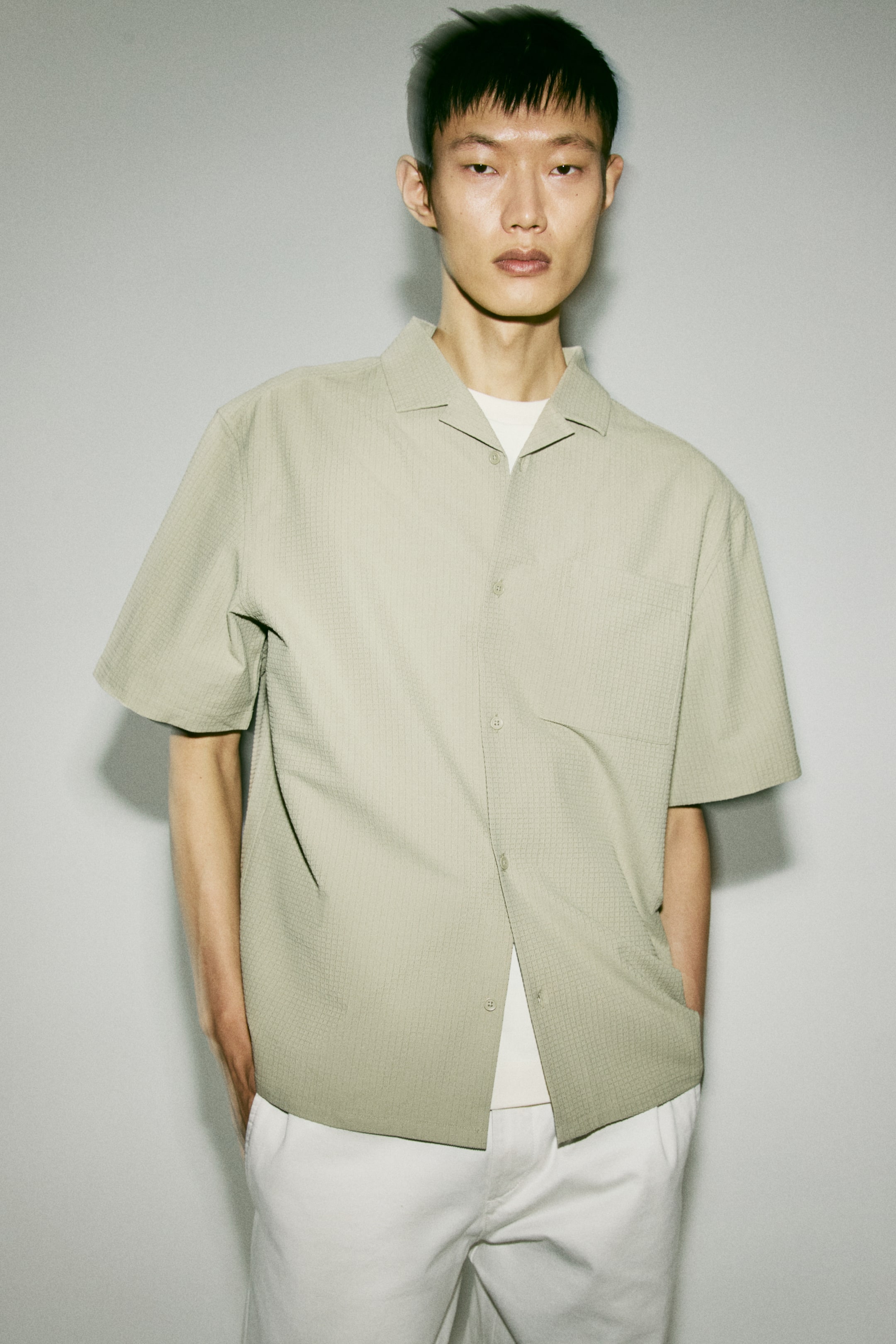 View larger image: A light green short-sleeved shirt featuring a subtle grid texture, a camp collar and a front button closure. It has a single patch pocket on the left chest, with a white T-shirt visible underneath.