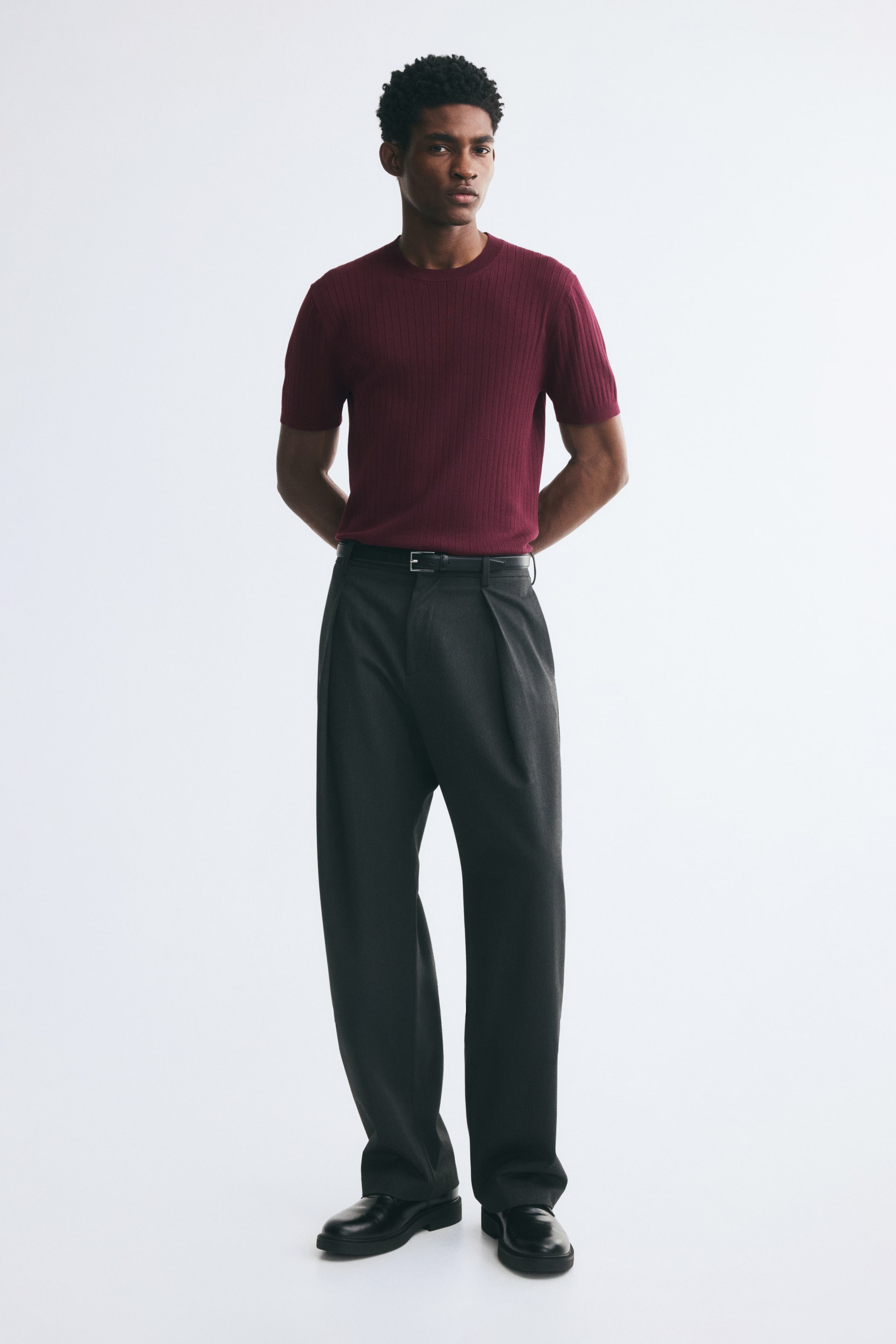 Voir une image plus grande: Un t-shirt côtelé fin bordeaux à coupe ajustée et à encolure ronde est porté par le mannequin. Le t-shirt à manches courtes est rentré dans un pantalon plissé gris foncé, porté avec une ceinture noire.