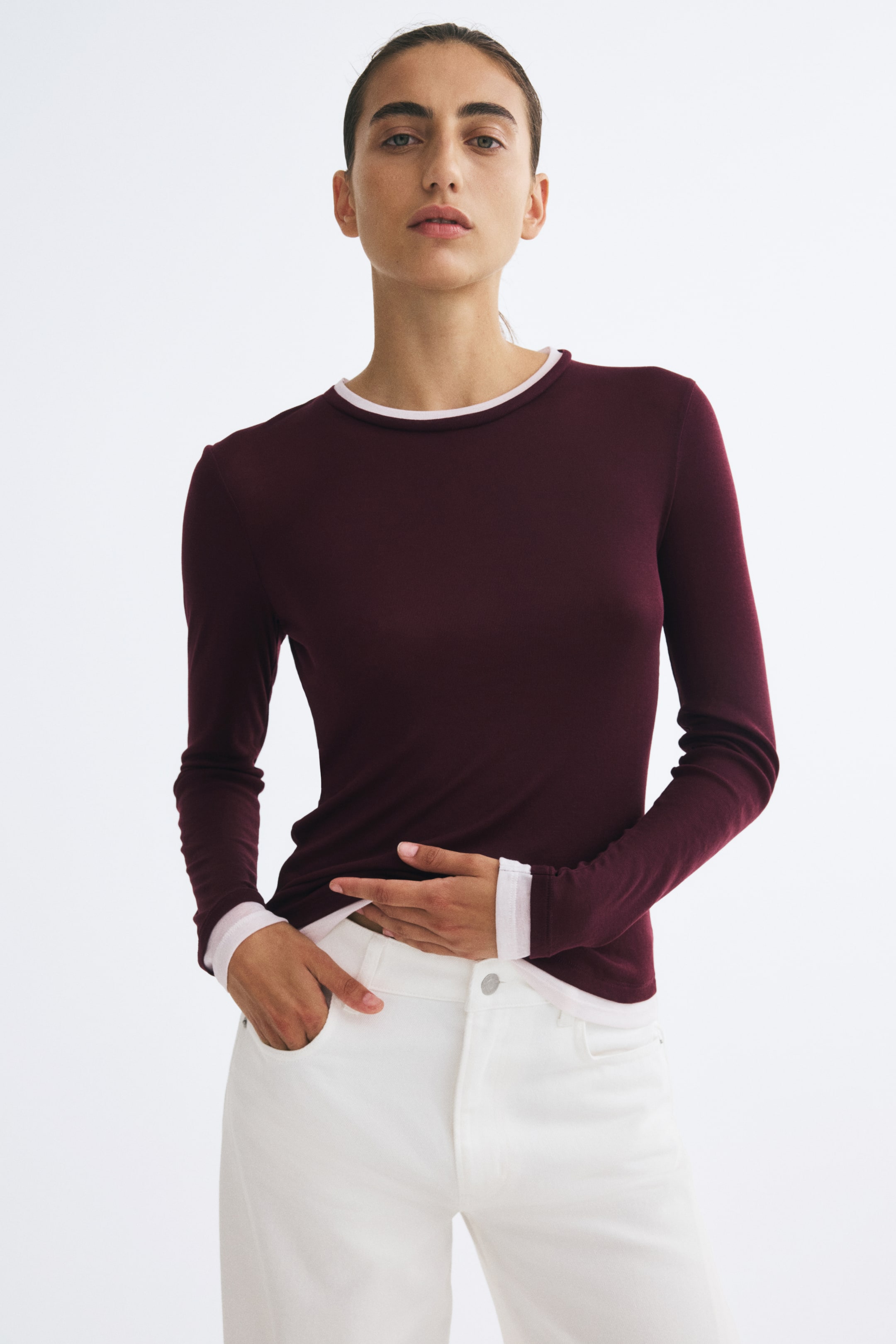 Ampliar la imagen: La modelo lleva puesta una blusa de punto fino de manga larga en color borgoña oscuro, con ribetes decorativos en rosa claro en el escote, los puños y el dobladillo. La modelo combina la blusa con pantalones blancos, apoyando una mano sobre la parte baja del torso y colocando la otra dentro del bolsillo.