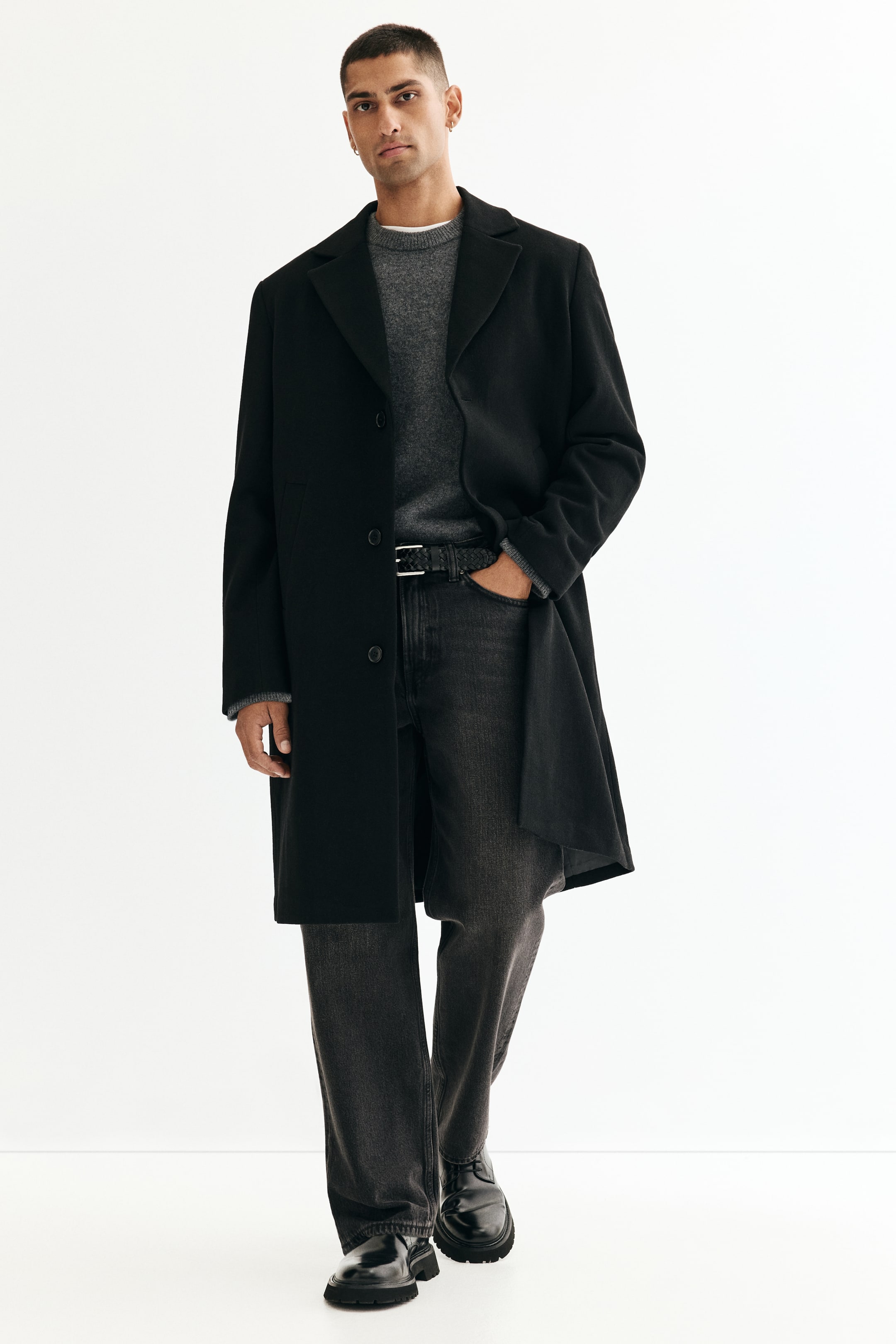 View larger image: A black single-breasted coat is being presented from the front, featuring notch lapels and a two-button front closure. The coat is extending to mid-thigh, with its long sleeves revealing the ribbed cuffs of a grey sweater.