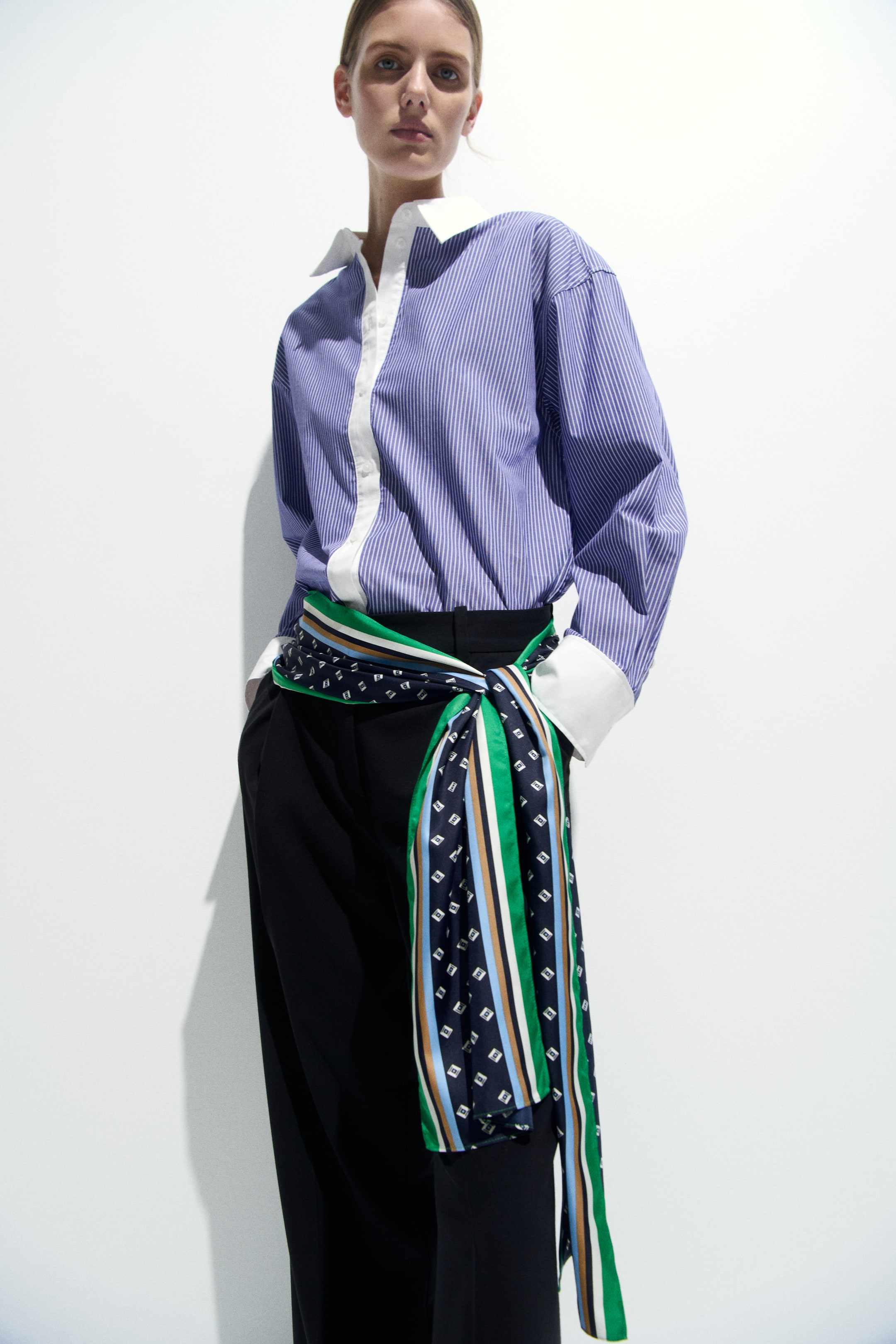 Größeres Bild ansehen: Ein blau-weiß gestreiftes Hemd in Oversized-Passform mit weißem Kragen und weißer Knopfleiste wird zu schwarzen Wide‑Leg‑Hosen kombiniert. Ein mehrgemusterter Schal mit dunkelblauen Bereichen mit weißen Quadraten und vertikalen Streifen in Grün, Weiß, Hellblau und Braun ist um die Taille gebunden, und die Enden hängen vorne herunter.