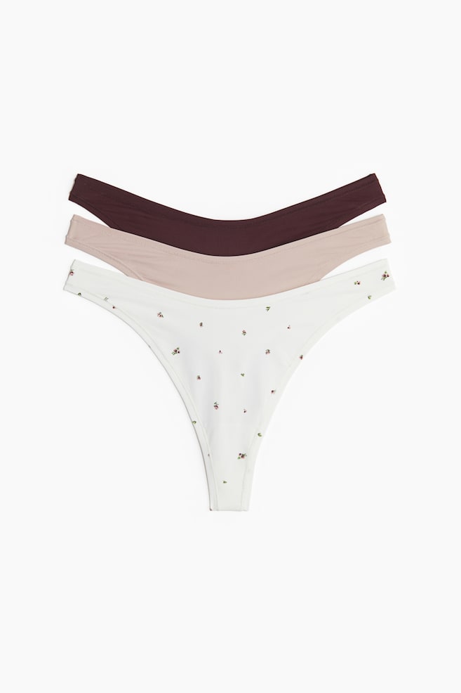 Three microfibre thong briefs are being displayed from the FRONT, each showing a low, V-shaped waist. The top brief is dark brown, the middle is light beige, and the bottom white brief is featuring a scattered small floral print. - 1