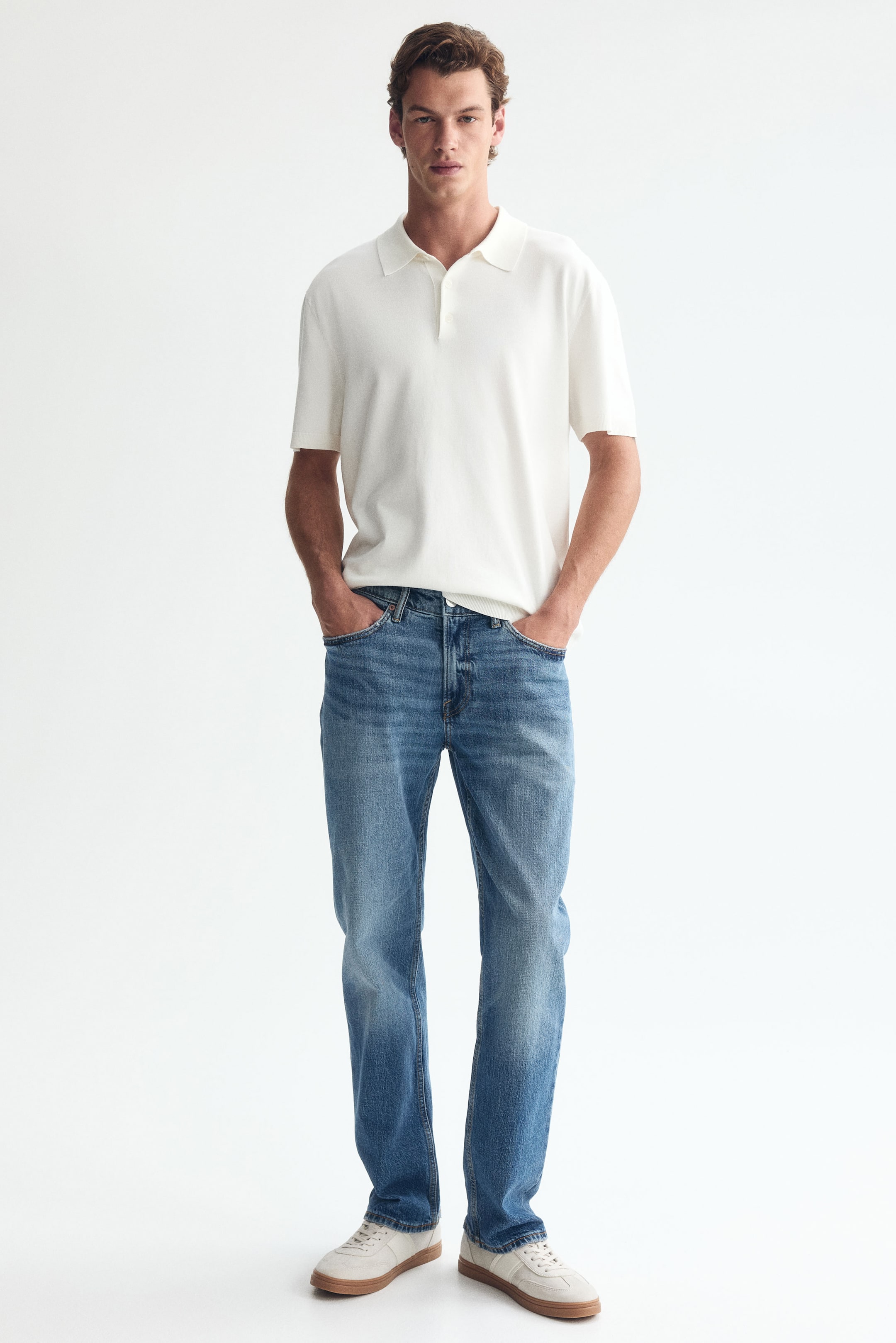 View larger image: Blue straight-leg jeans in a medium wash are being worn by the model, who is standing with hands in the front pockets. The jeans feature a regular fit and are styled with a white short-sleeved polo shirt and light-coloured sneakers.