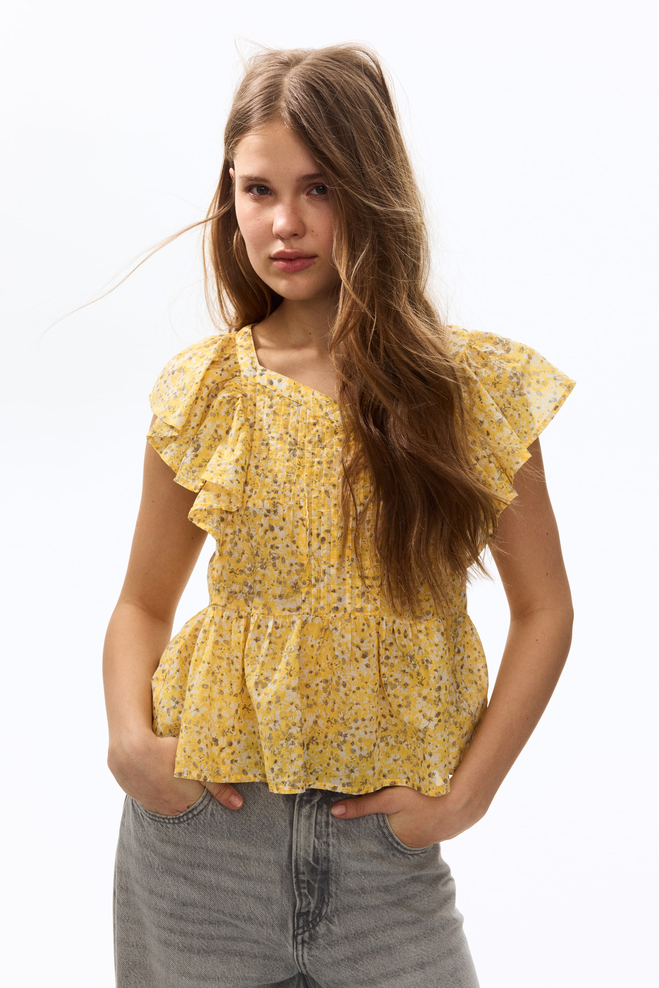 Büyük resmi görüntüle: Model, hafif pamuklu dokumada, küçük çiçek desenli, ön kısmında pilikaşeler bulunan, kalp yaka, omuzlardan devam eden fırfır detaylı ve bel kısmında toplanarak hafifçe genişleyen peplum oluşturan sarı kolsuz bir üst giymektedir. Model ayrıca gri kot pantolon giymiş olup, elleri pantolonun ceplerinde durmaktadır.