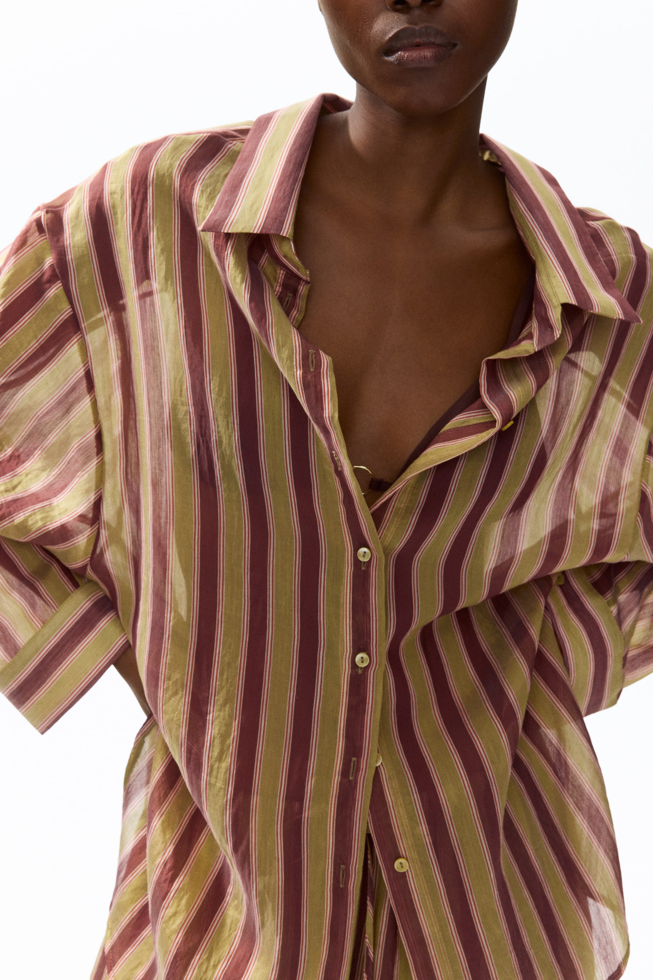 Visualizza immagine più grande: Una camicia con bottoni presenta strisce verticali in bordeaux e verde oliva, con un colletto aperto a punta e bottoni chiari lungo la parte anteriore. Il tessuto leggero e leggermente trasparente è indossato sbottonato in alto, rivelando una spallina del reggiseno sottostante.