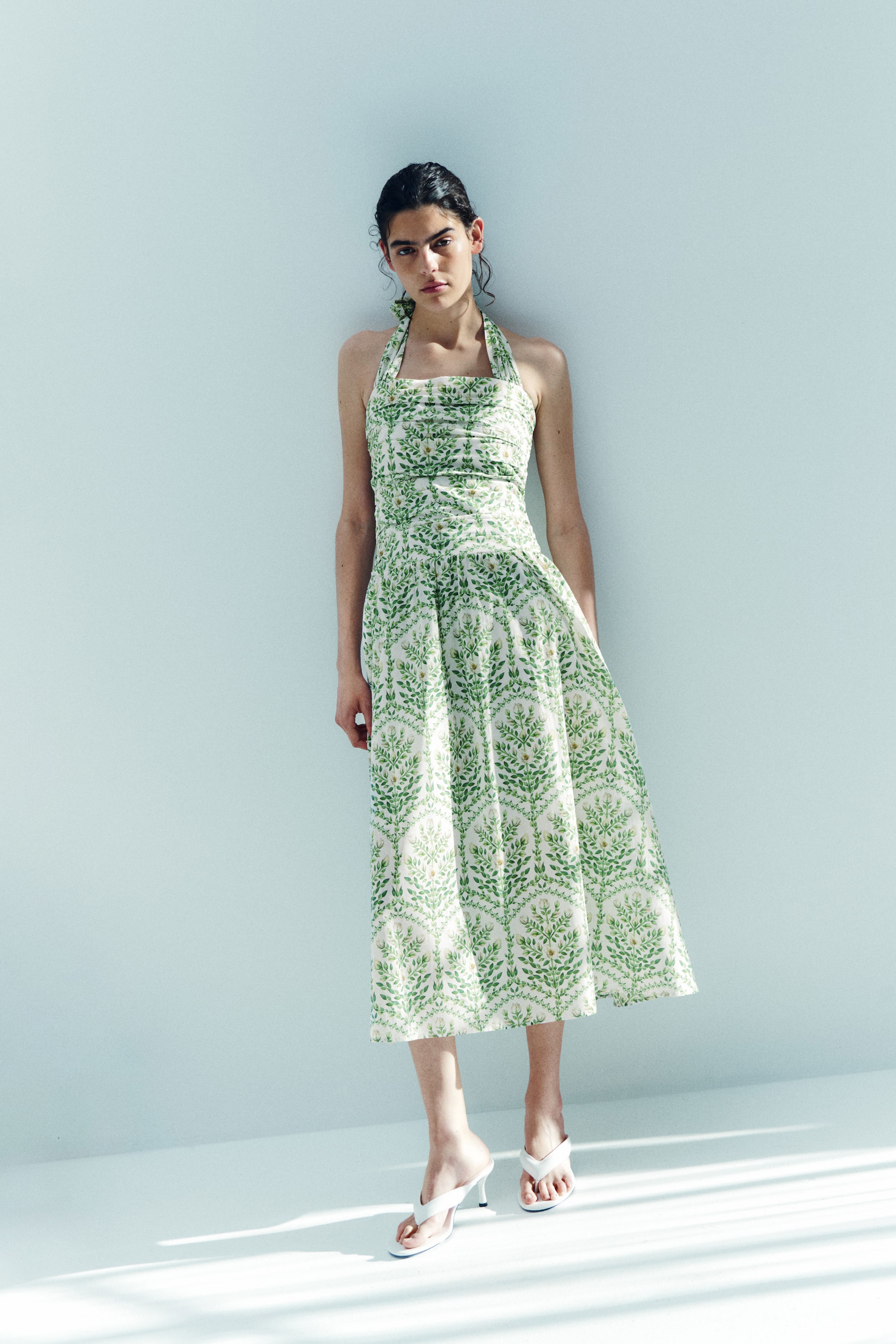 View larger image: A white halter neck midi dress with a green leaf pattern features a ruched bodice and a flowing A-line skirt. It is shown front-facing and styled with white high-heeled thong sandals.