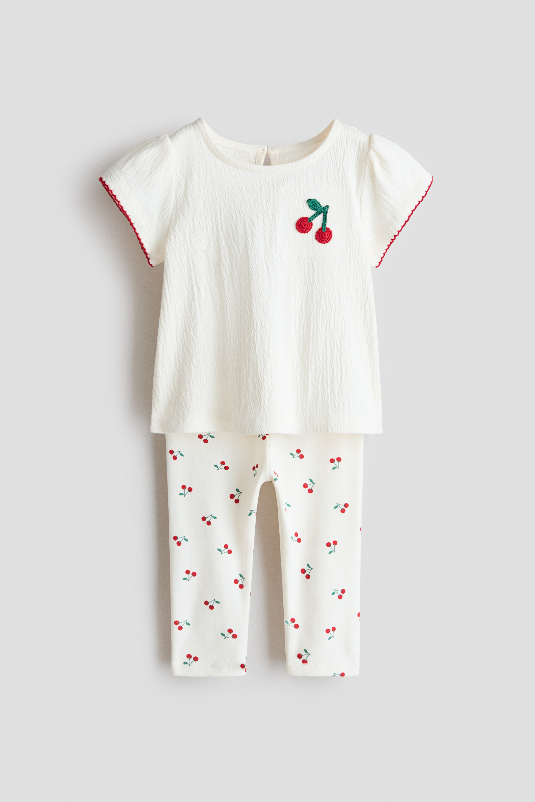 View larger image: A two-piece baby outfit is shown front-facing, featuring an off-white, short-sleeved top with a crinkled texture, red scalloped trim on the sleeves, and an embroidered cherry motif on the left chest. The matching off-white leggings are patterned with small red cherries with green stems.