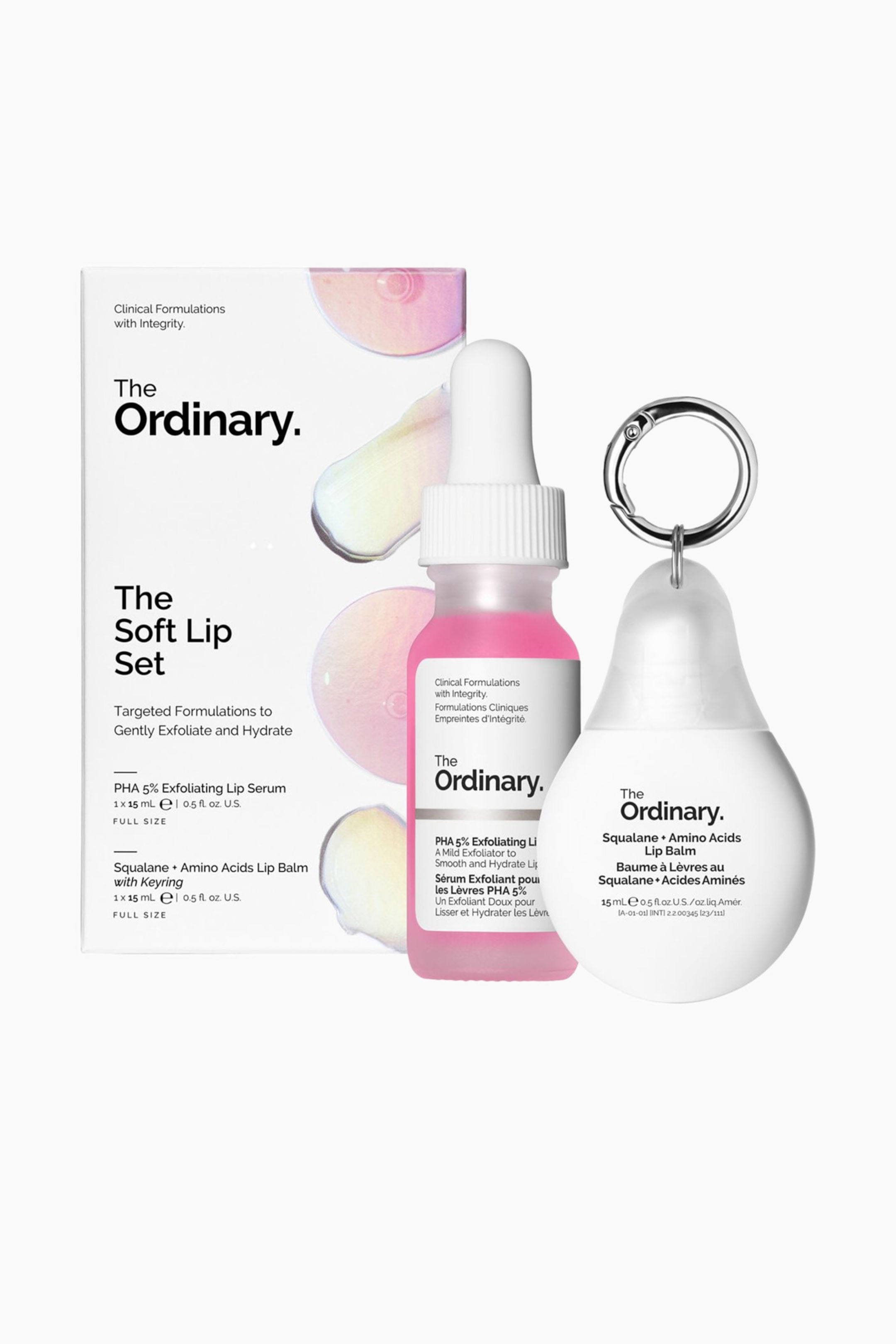 Vis større bilde: Soft Lip Set fra The Ordinary vises med den hvite, rektangulære emballasjeboksen stående bak de to hovedproduktene. I forgrunnen står en gjennomsiktig pipetteflaske med et knallrosa eksfolierende serum ved siden av en hvit leppepomadebeholder formet som en dråpe og med en sølvfarget metallnøkkelring.