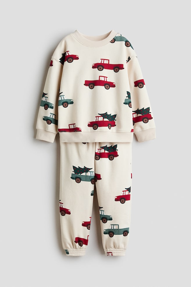 A cream-coloured fleece sweater and track pants set is shown from the front, featuring an all-over print of red and green pickup trucks carrying pine trees. The long-sleeved sweater features a ribbed crew neck, dropped shoulders and ribbed trims, while the matching pants feature elasticated ankle cuffs. - 1
