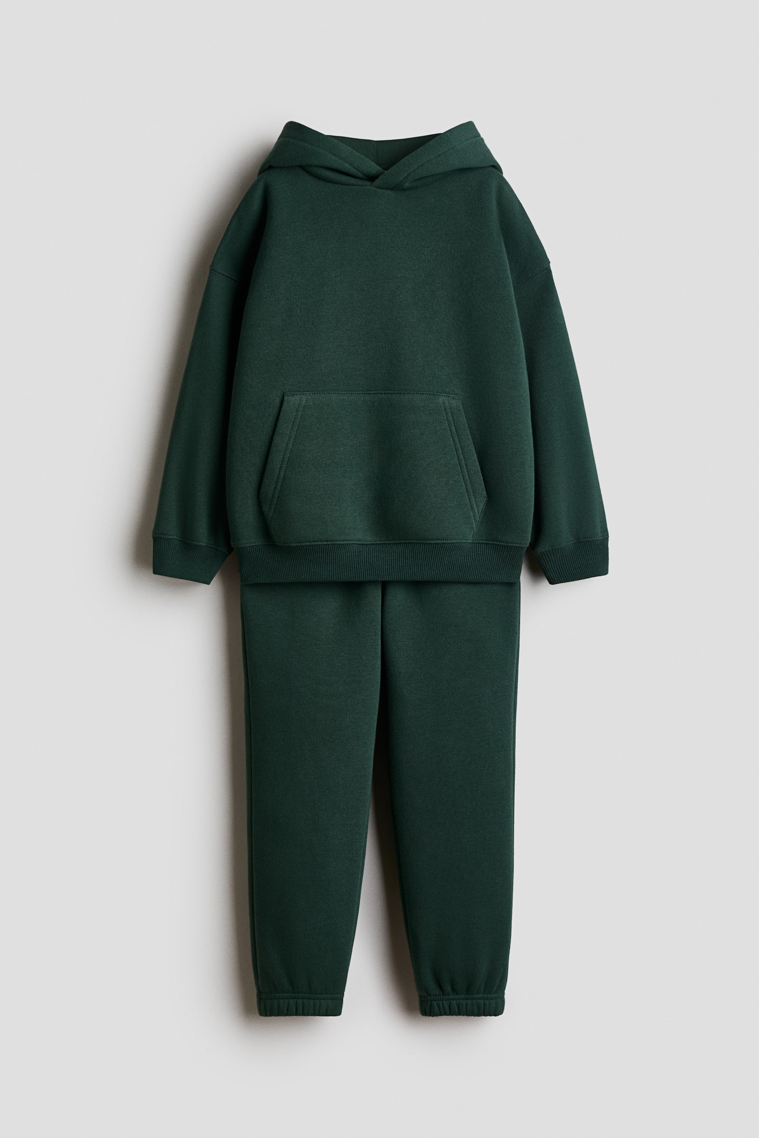 A dark green tracksuit is shown from the front, featuring a long-sleeved hoodie with a front kangaroo pocket, ribbed cuffs, and a ribbed hem. The matching sweatpants are displayed directly below, with elasticated cuffs at the ankles.