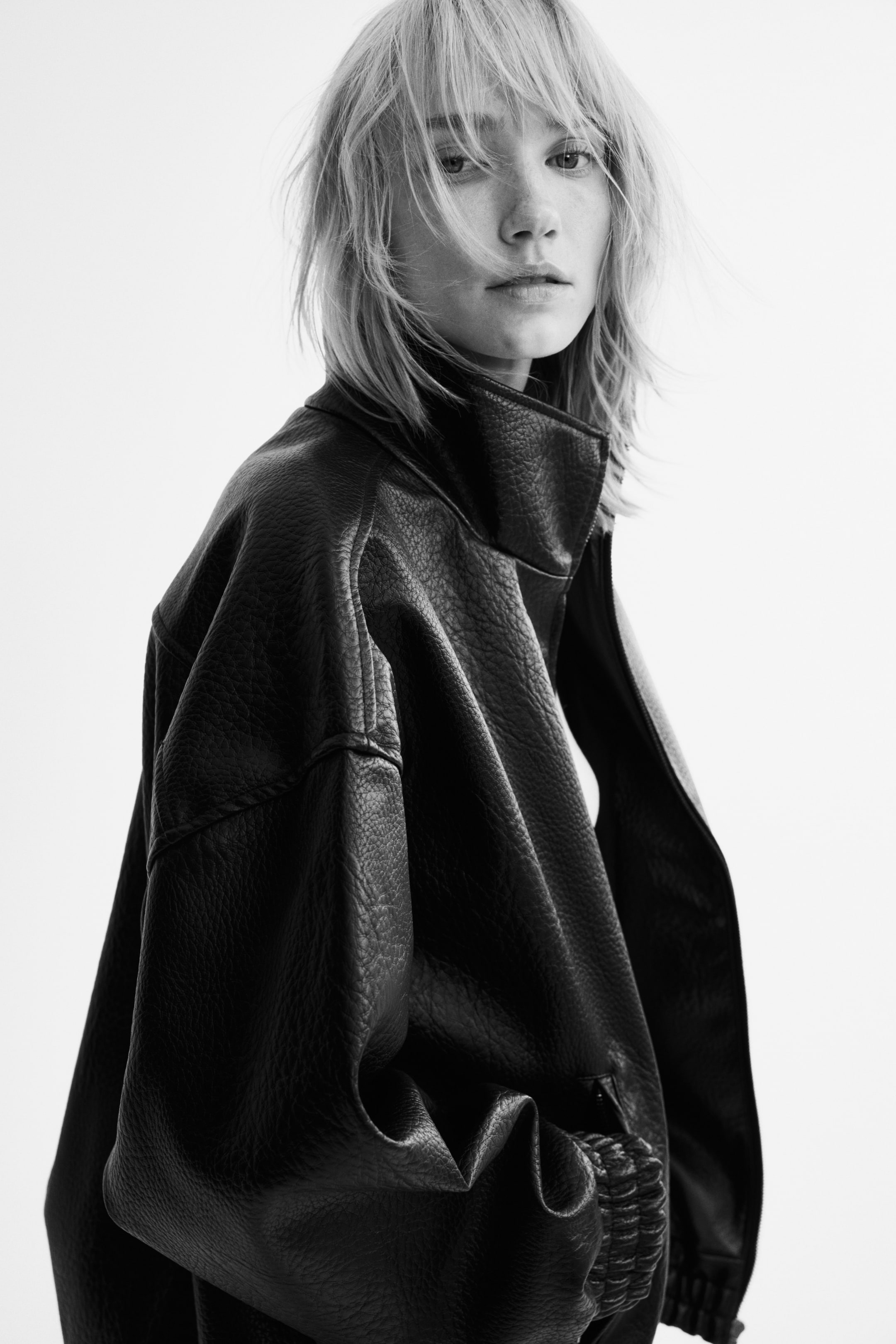 Ampliar la imagen: La modelo lleva puesta una chaqueta oversize en una tela recubierta oscura y arrugada. La chaqueta presenta cuello alto tipo mao, hombros caídos y puños con elástico fruncido.