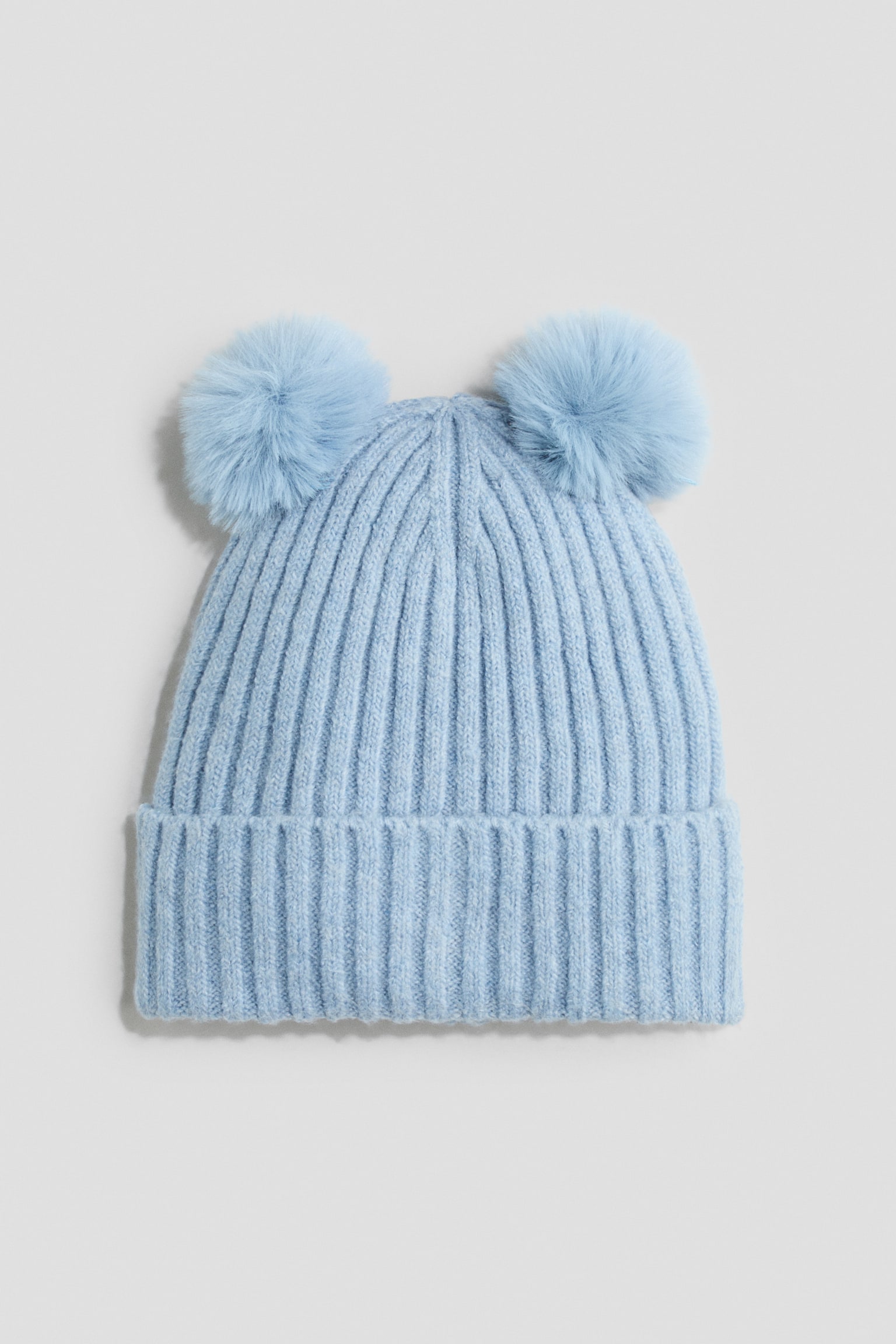 A light blue ribbed knit beanie is shown from the front, featuring a thick folded cuff and two matching fluffy pom-poms attached to the top corners.