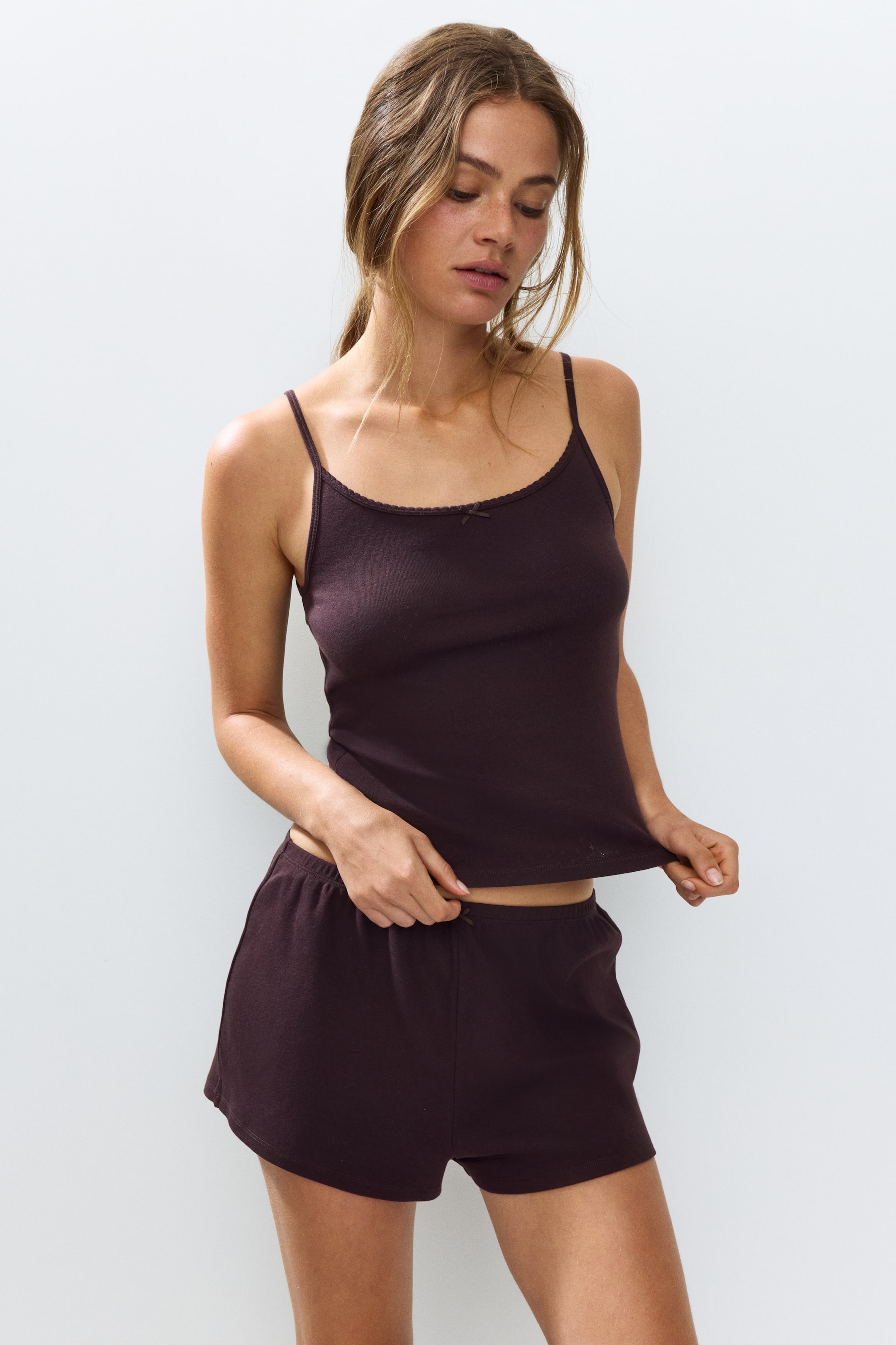 View larger image: A dark brown camisole top and matching shorts set is showcased from a front angle. The camisole features thin spaghetti straps, a scalloped trim along the neckline, and a small bow at the center front. The shorts display an elastic waistband, and the fabric of both pieces is gently pulled to demonstrate its texture and fit.
