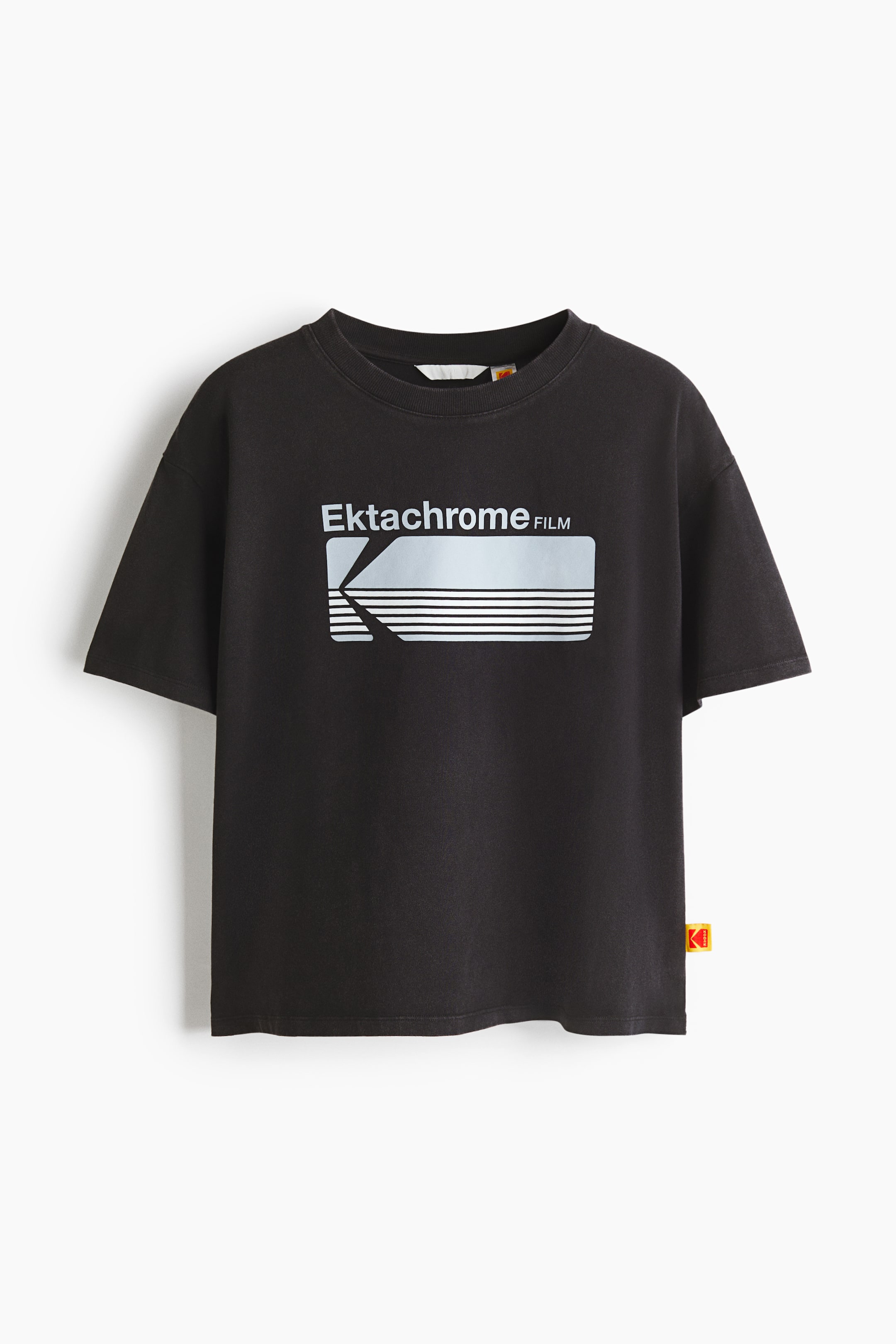 View larger image: A black short-sleeved t-shirt is displayed from the front, featuring a crew neck. A large white graphic is centred on the chest, showcasing "Ektachrome FILM" text above a stylised "K" logo with horizontal lines. A small red and yellow tag is visible on the lower right side seam.