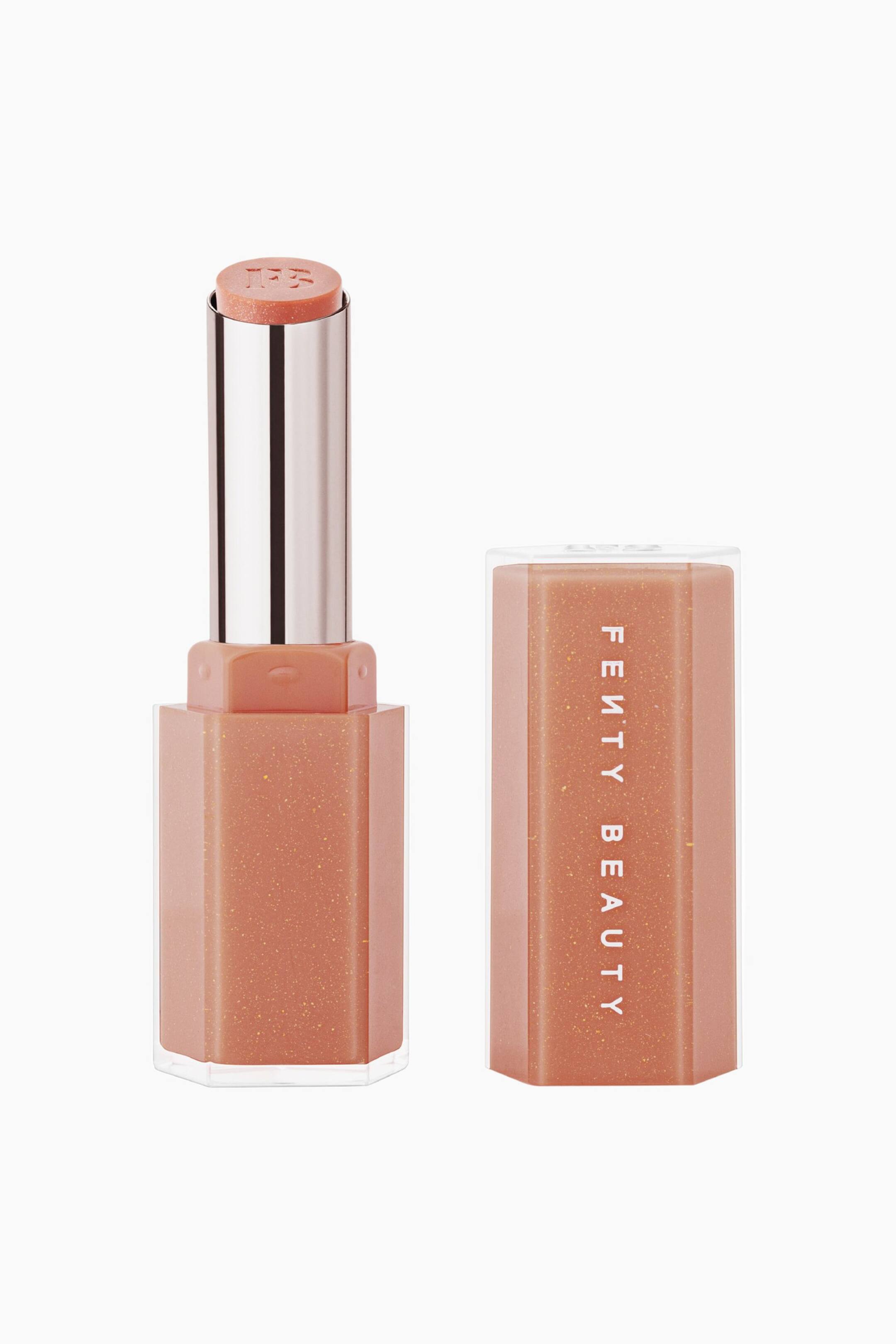 Visa större bild: Gloss Bomb Stix High-shine Shimmer Gloss Stick står upprätt med korken av, och visar den persikoskimmrande gloss-stiftet utdraget ur ett silverfärgat metalliskt innerhylsa. Den matchande sexkantiga, genomskinliga capsen i persikobeige står intill och visar "FENTY BEAUTY" tryckt vertikalt i vitt.