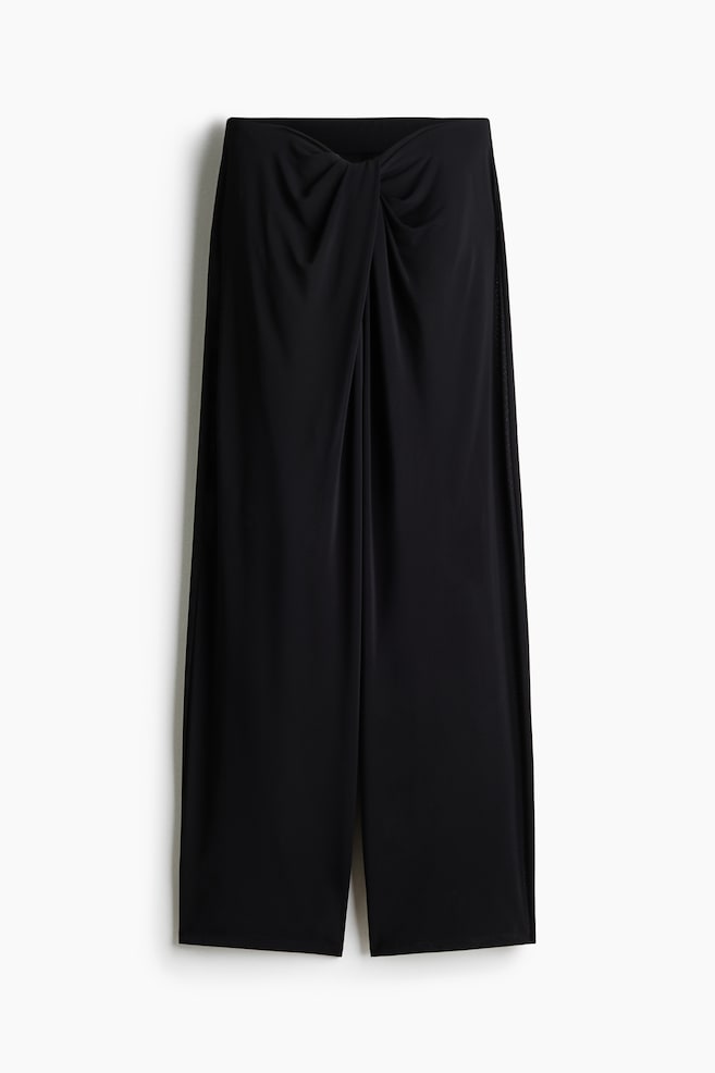 Black wide-leg pants are showcased from the front, highlighting a striking twisted knot detail at the center front of the waistband. The sleek, black fabric drapes into a relaxed, flowing silhouette. - 1