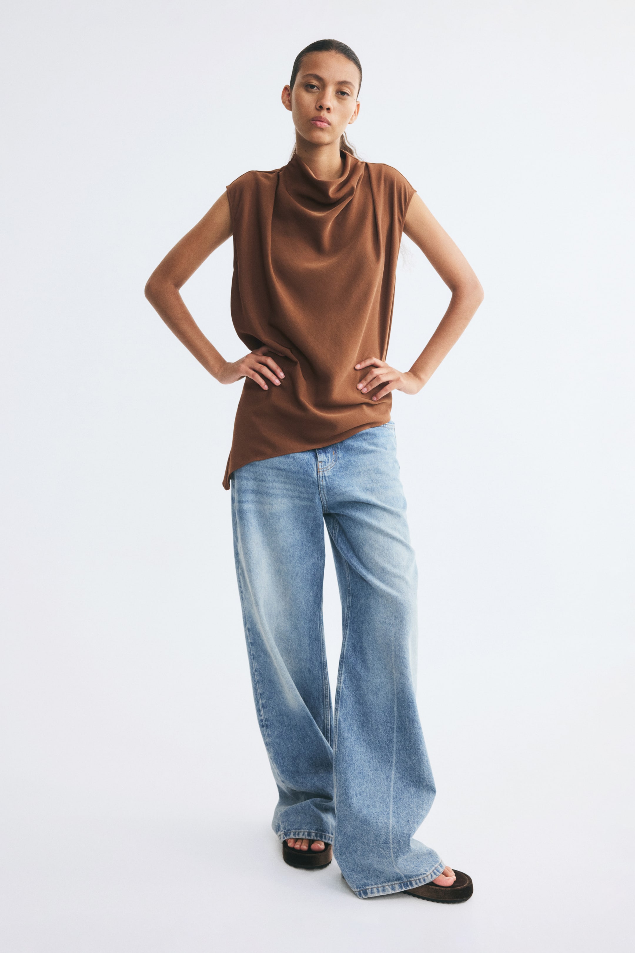 View larger image: The model is wearing a brown sleeveless top made from woven fabric, featuring a draped cowl neckline and an asymmetrical hem. It is styled with wide-leg, light blue denim jeans.