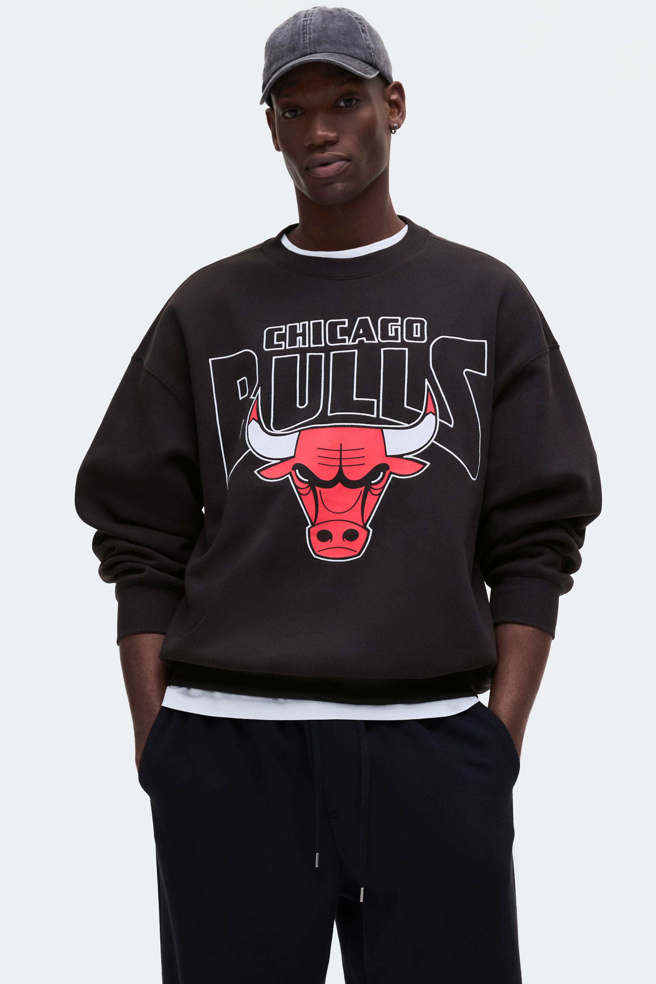 Visa större bild: En svart oversized sweatshirt med rund hals har en "Chicago Bulls"-logotyp med vit och svart kantad text och en röd och vit tjurhuvudgrafik på framsidan. Den har ribbade muddar och en delvis synlig ribbad fåll, med en vit t-shirt synlig under dess halslinje och nederkant.
