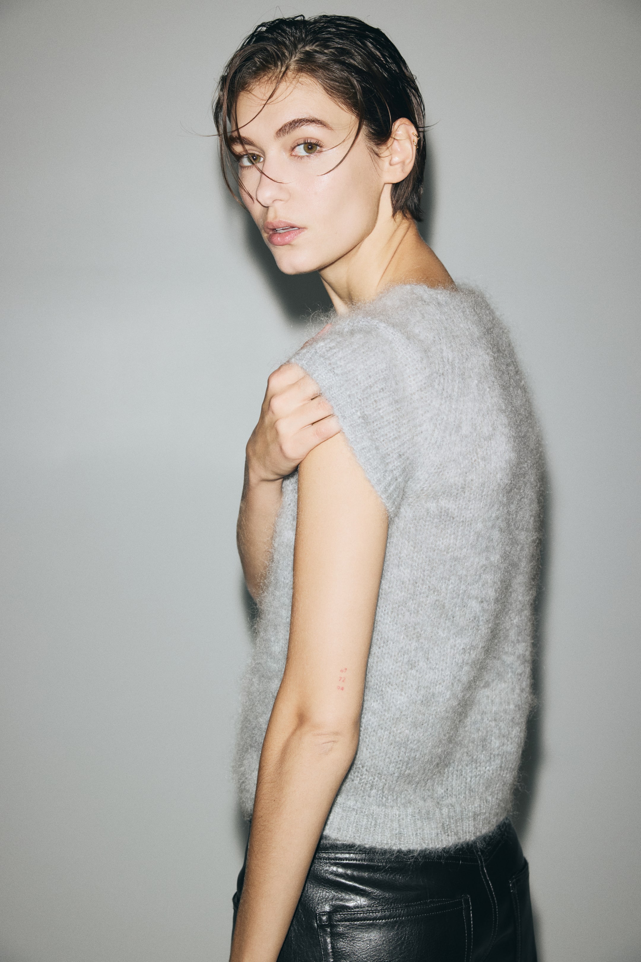 Ampliar la imagen: Se muestra un top sin mangas de punto color gris claro con textura peluda desde un ángulo lateral-frontal. Presenta un ajuste holgado y un dobladillo acanalado, y se lleva con pantalones de cuero negros.