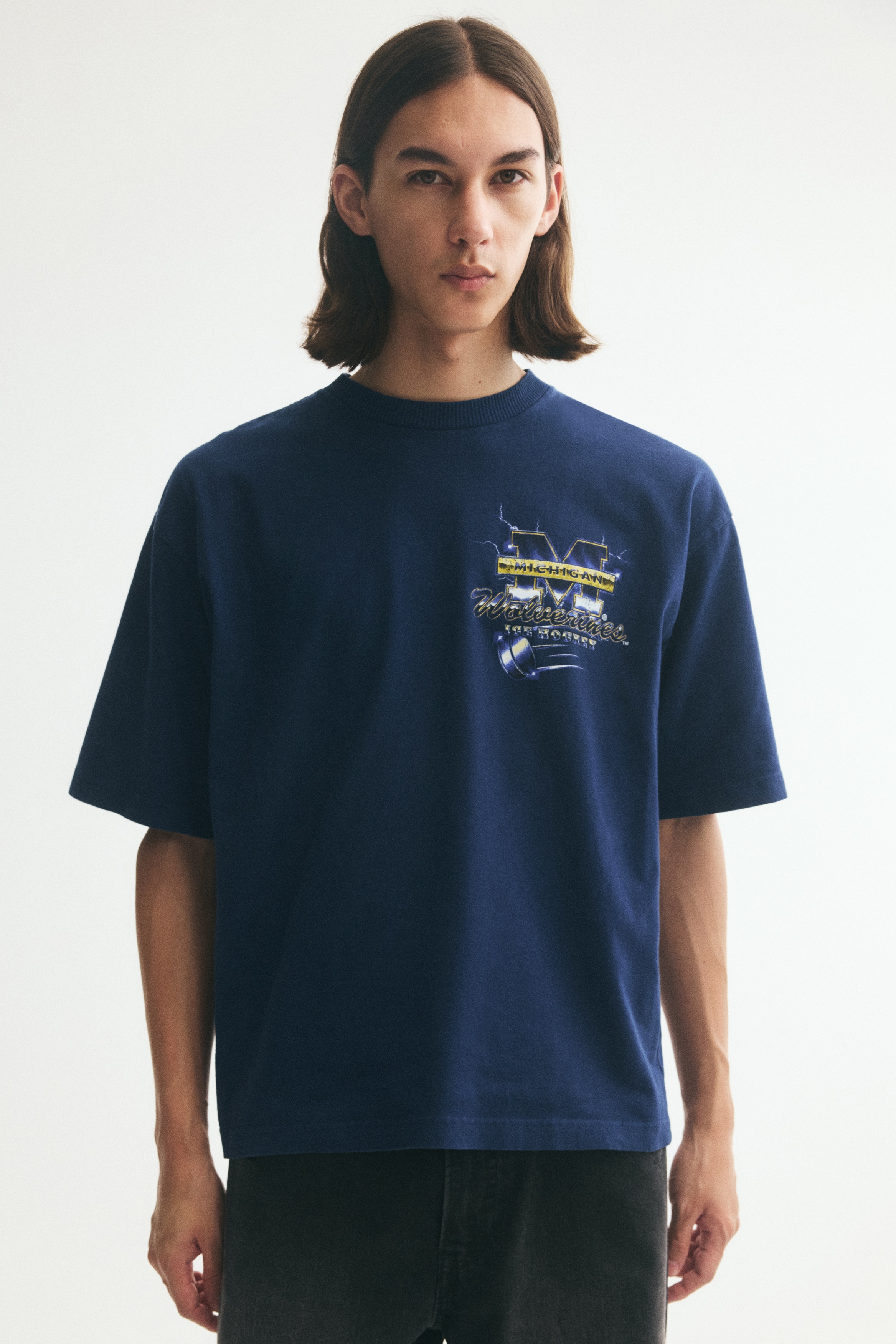 A navy blue heavyweight cotton jersey T-shirt is hanging in a front-facing view, featuring a relaxed fit with a ribbed crew neck, dropped shoulders, and a straight hem. The T-shirt is displaying a graphic print of "Michigan Wolverines Ice Hockey" on the left chest.