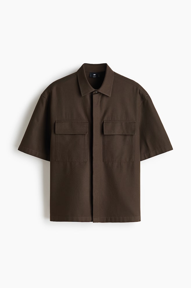 A dark brown, short-sleeved overshirt is facing the front, featuring a classic collar and a concealed button placket. The shirt is displaying two large chest pockets with flaps and wide, relaxed-fit sleeves. - 1