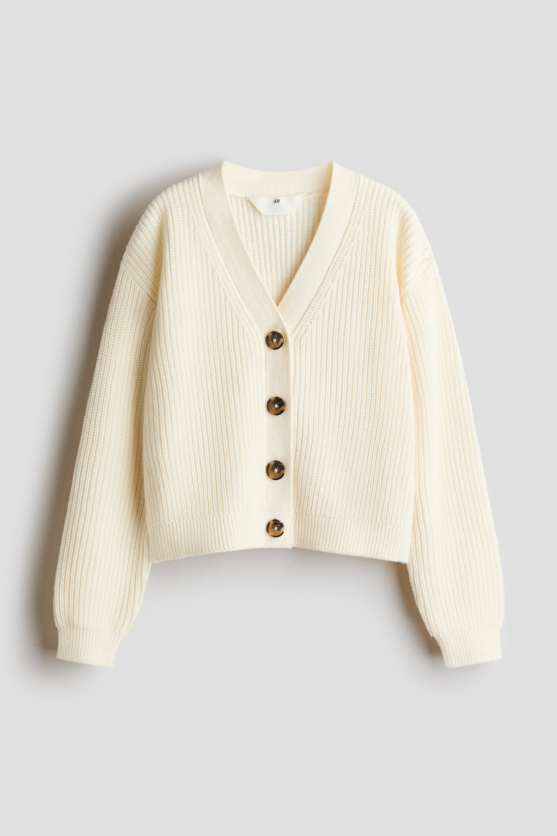 View larger image: A cream cardigan is presenting its FRONT, featuring a V-shaped neckline and five brown buttons down the center. It is showcasing a soft rib knit texture with dropped shoulders and ribbing at the opening, cuffs, and hem.