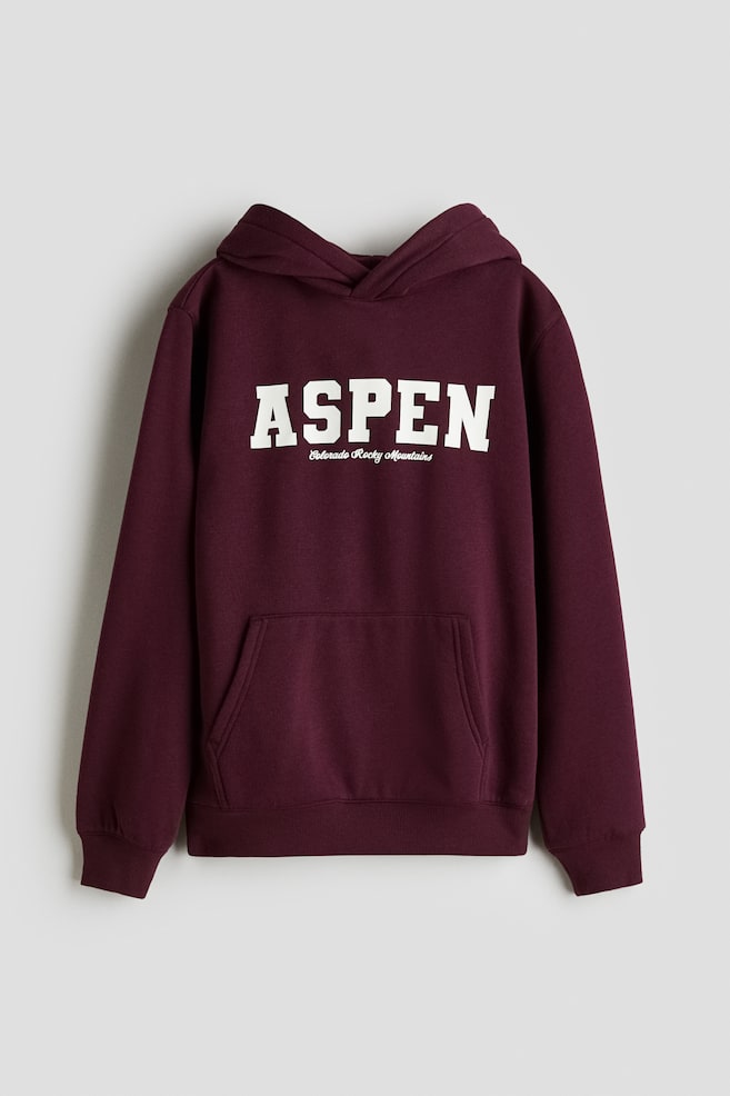 A dark red cotton sweatshirt hoodie is displayed from the front, featuring "ASPEN" in white block letters with "Colorado Rocky Mountains" in smaller script below. It includes a lined hood with a wrap front, dropped shoulders, a kangaroo pocket, ribbed cuffs, and a ribbed hem. - 1