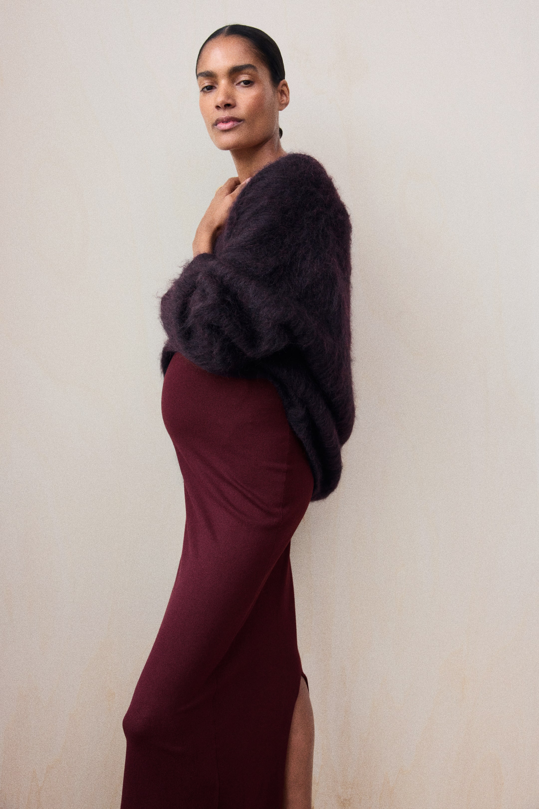 View larger image: The model is wearing an ankle-length, fitted skirt in burgundy fine knit. The high-waisted skirt features a slit at the back hem and is styled with a dark, textured fluffy sweater draped over the upper body.