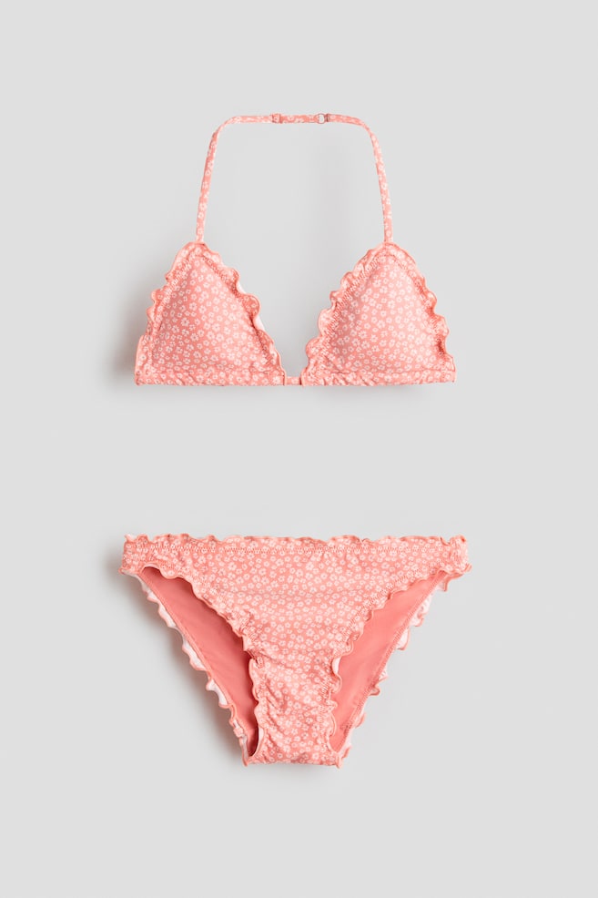 Ein Pfirsich-farbenes Bikini-Set mit Blumenmuster zeigt seine Vorderansicht. Das Triangel-Oberteil verfügt über gerüschte Kanten und einen verstellbaren Neckholder-Träger mit Hakenverschluss, während der passende Slip mit gerüschten Kanten und gerafften Details an der Vorderseite versehen ist. - 1
