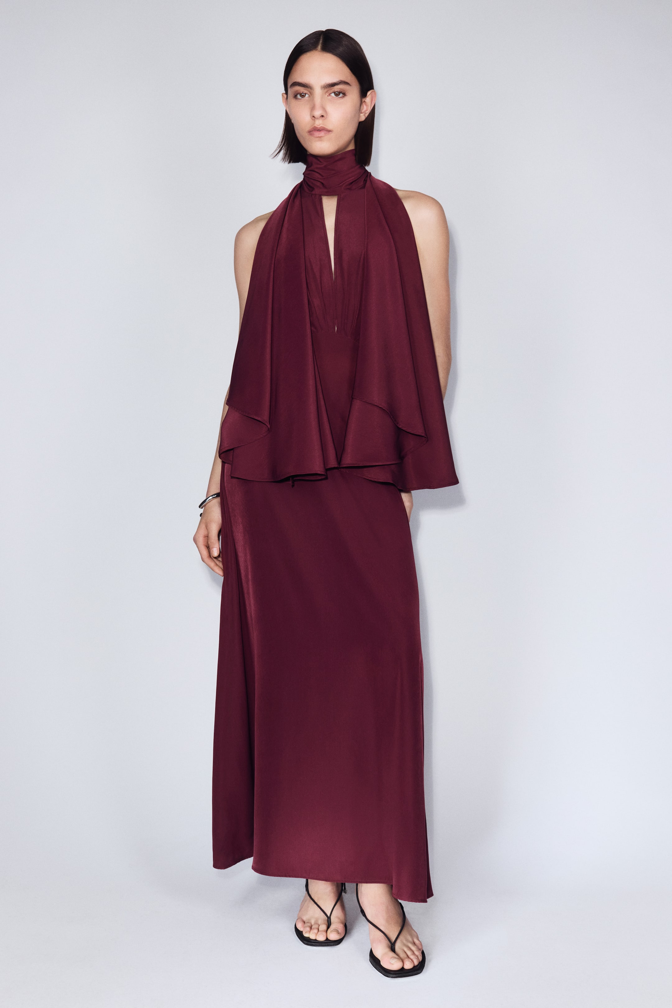 Ampliar la imagen: La modelo lleva puesto un vestido largo de color rojo intenso tejido. El vestido presenta un cuello alto con una abertura frontal en forma de V y mangas tipo capa voluminosas que se extienden hacia la espalda, creando un efecto drapeado sobre la parte superior del cuerpo, con un dobladillo de corte recto.