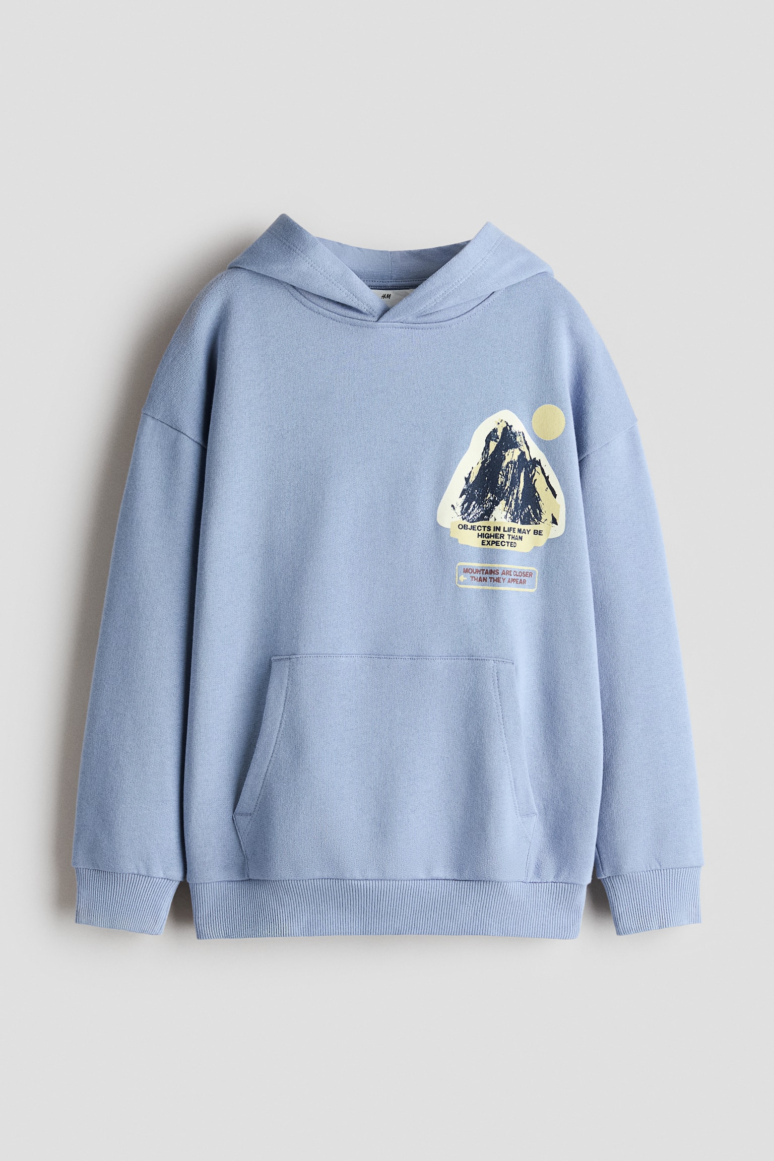 A light blue hoodie is shown from the front, featuring a graphic print of a mountain peak on the left chest with the text "OBJECTS IN LIFE MAY BE HIGHER THAN EXPECTED." The hoodie features dropped shoulders, a front kangaroo pocket, and ribbed trim on the cuffs and hem.
