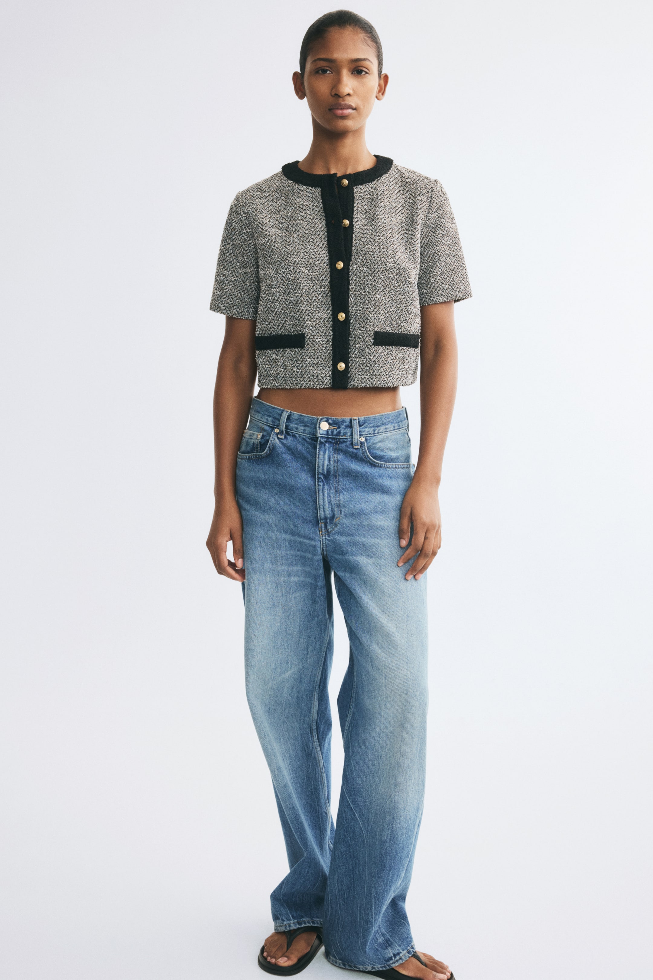 Xem ảnh lớn hơn: Một chiếc áo khoác dệt kim tay ngắn dáng crop với họa tiết tweed dệt nổi màu xám đang được người mẫu mặc. Áo khoác có viền tương phản màu đen dọc theo cổ tròn, nẹp giữa thân và túi phía trước, được cài bằng nút tông vàng và phối cùng quần jeans xanh ống rộng.