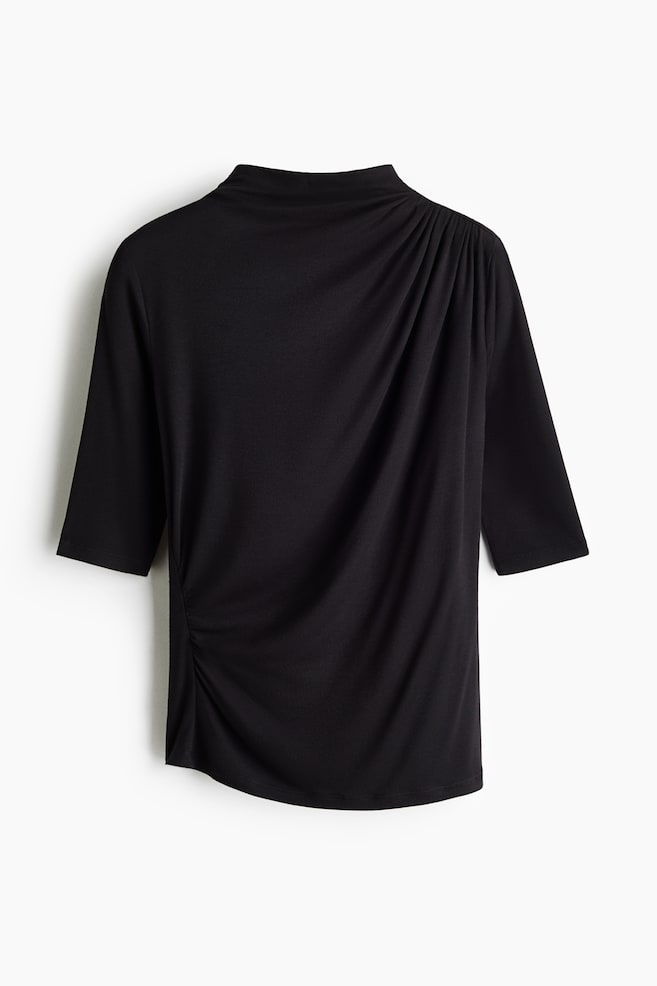 This black, elbow-sleeve top is shown from the front, featuring a high neckline with gathers at the left shoulder that create an elegant diagonal draping across the body. The draping flows down to a ruched detail at the right side seam, creating a subtly asymmetrical hemline. - 1