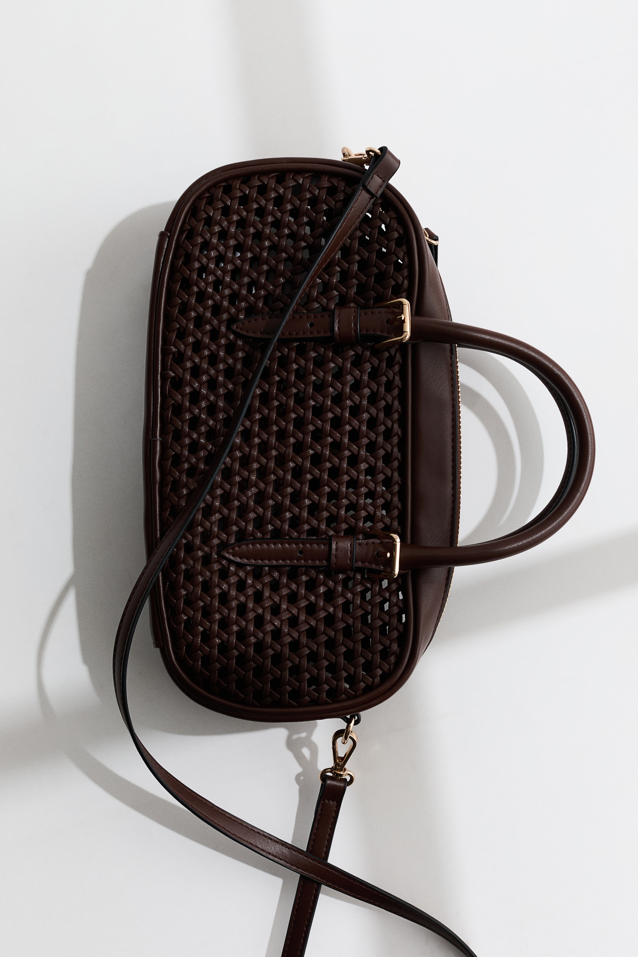 View larger image: A dark brown oval handbag is lying flat, its top and bottom panels showcasing a woven pattern with smooth sides. It features two top handles attached by buckled straps and a detachable shoulder strap, all accented with gold-toned hardware.