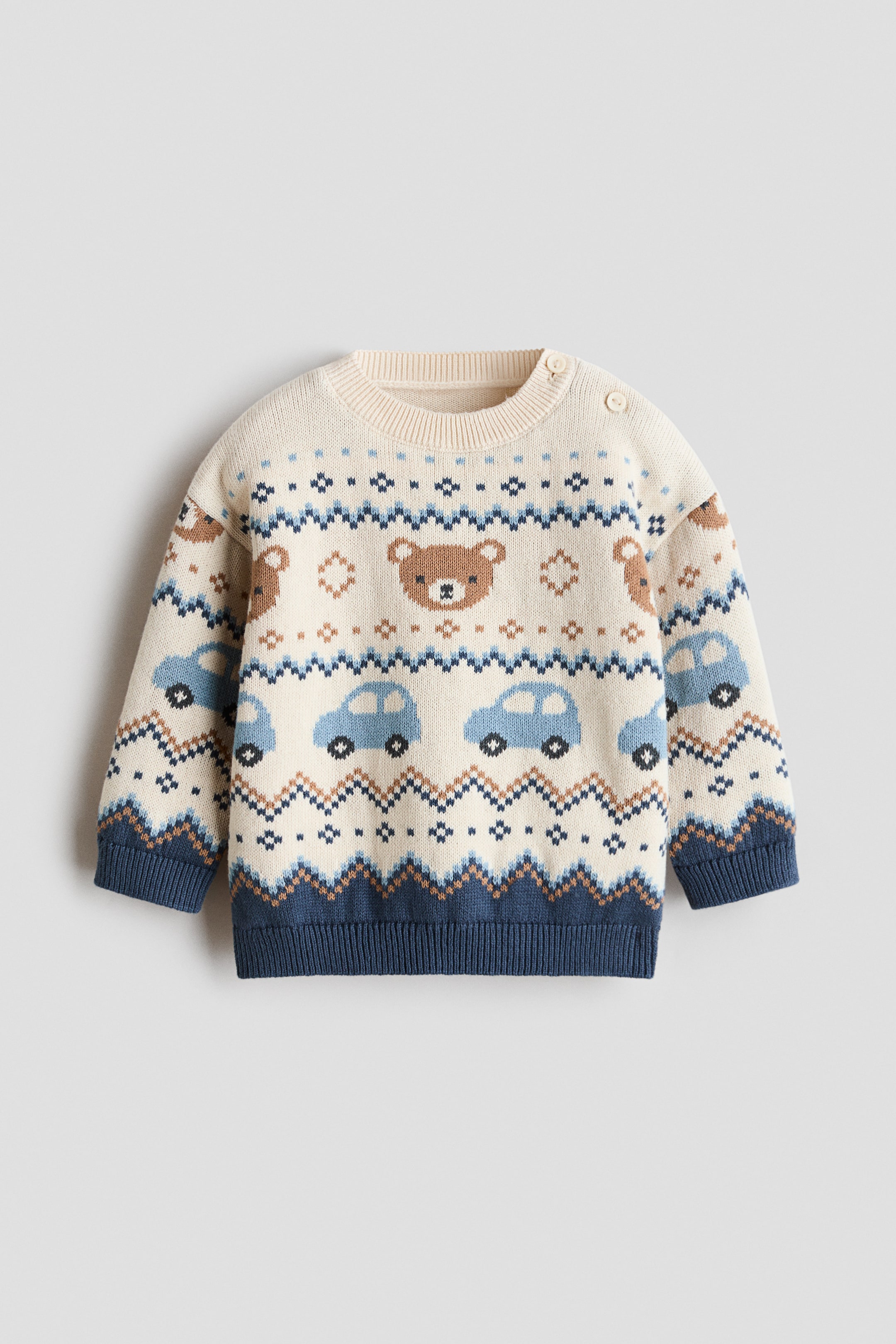 Agrandir l'image: Un pull crème pour enfants arbore un motif Fair Isle avec des visages d'ours bruns, des voitures bleu clair et des formes géométriques en brun, bleu clair et bleu foncé, visibles sur le devant. Il présente un col rond côtelé avec deux boutons sur l'épaule gauche, ainsi que des poignets et un ourlet côtelés bleu foncé.