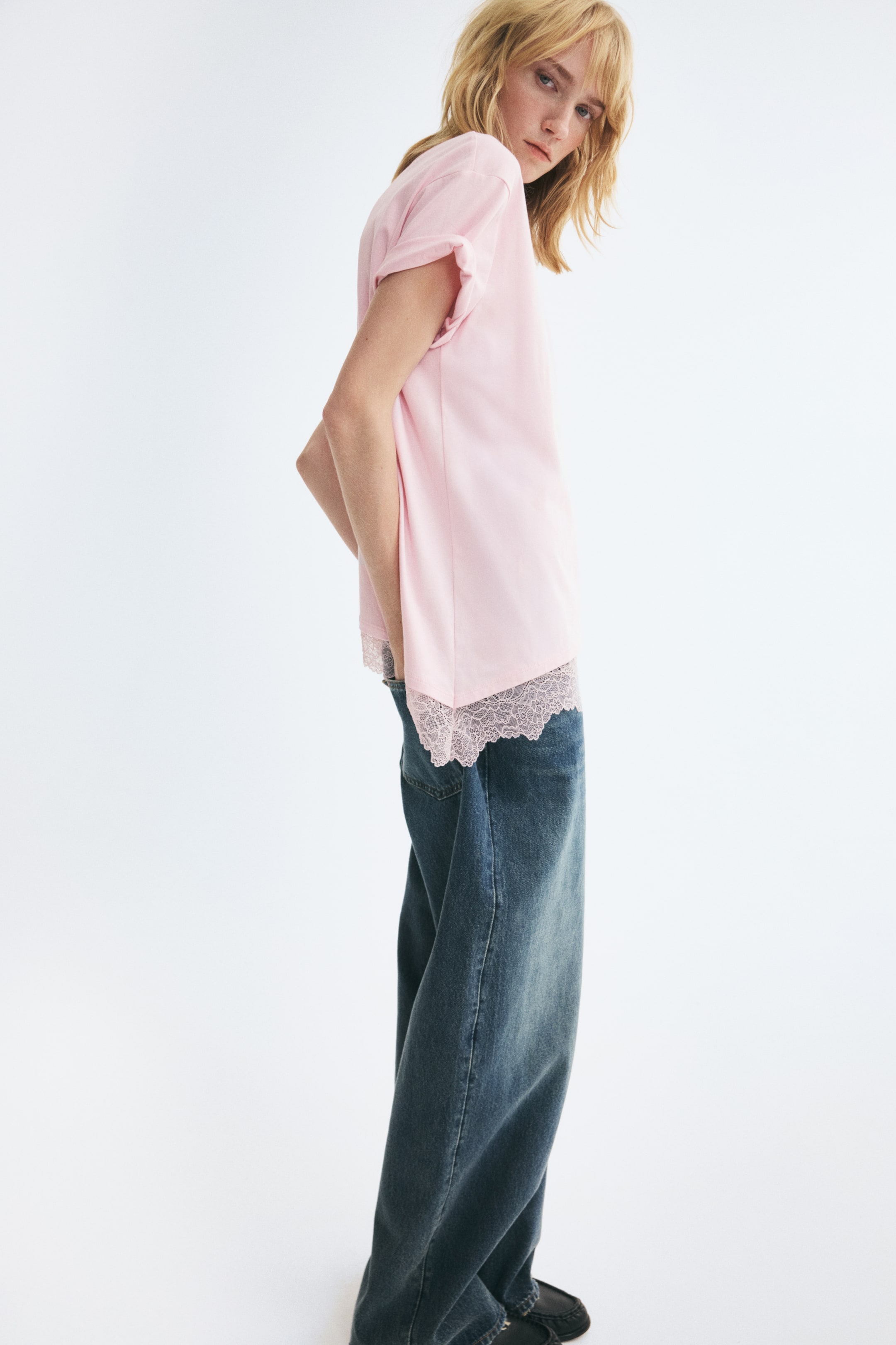 View larger image: A light pink relaxed-fit t-shirt featuring rolled short sleeves and an asymmetrical hem with scalloped lace trim is being worn by the model. The t-shirt is being styled with wide-leg blue denim jeans as the model is standing in profile.