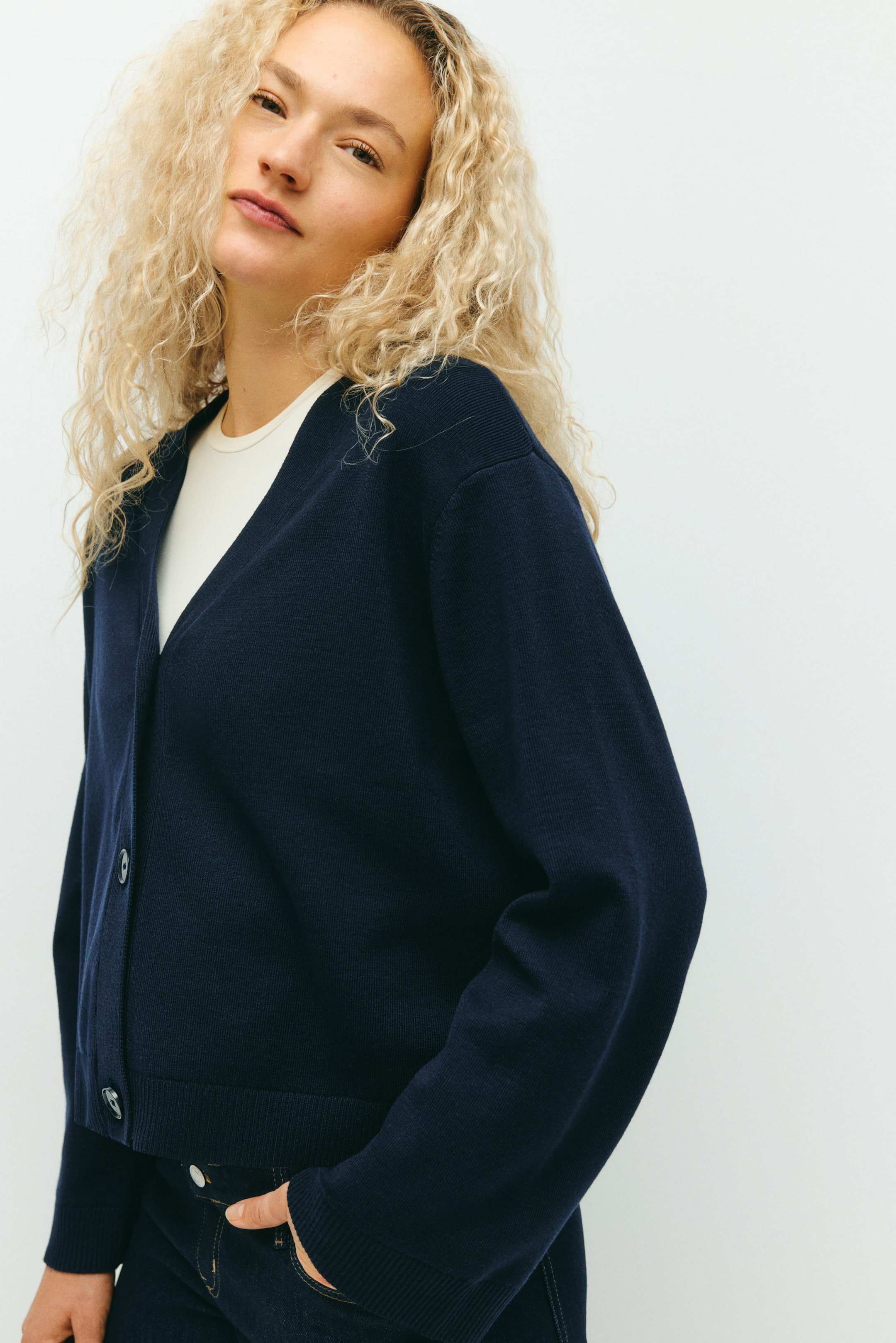 View larger image: A navy blue V-neck cardigan is being presented, angled slightly to the left, featuring a button-front closure with dark buttons. It is showing a relaxed fit with dropped shoulders, long sleeves, and a ribbed hem, being worn open over a light-colored top.