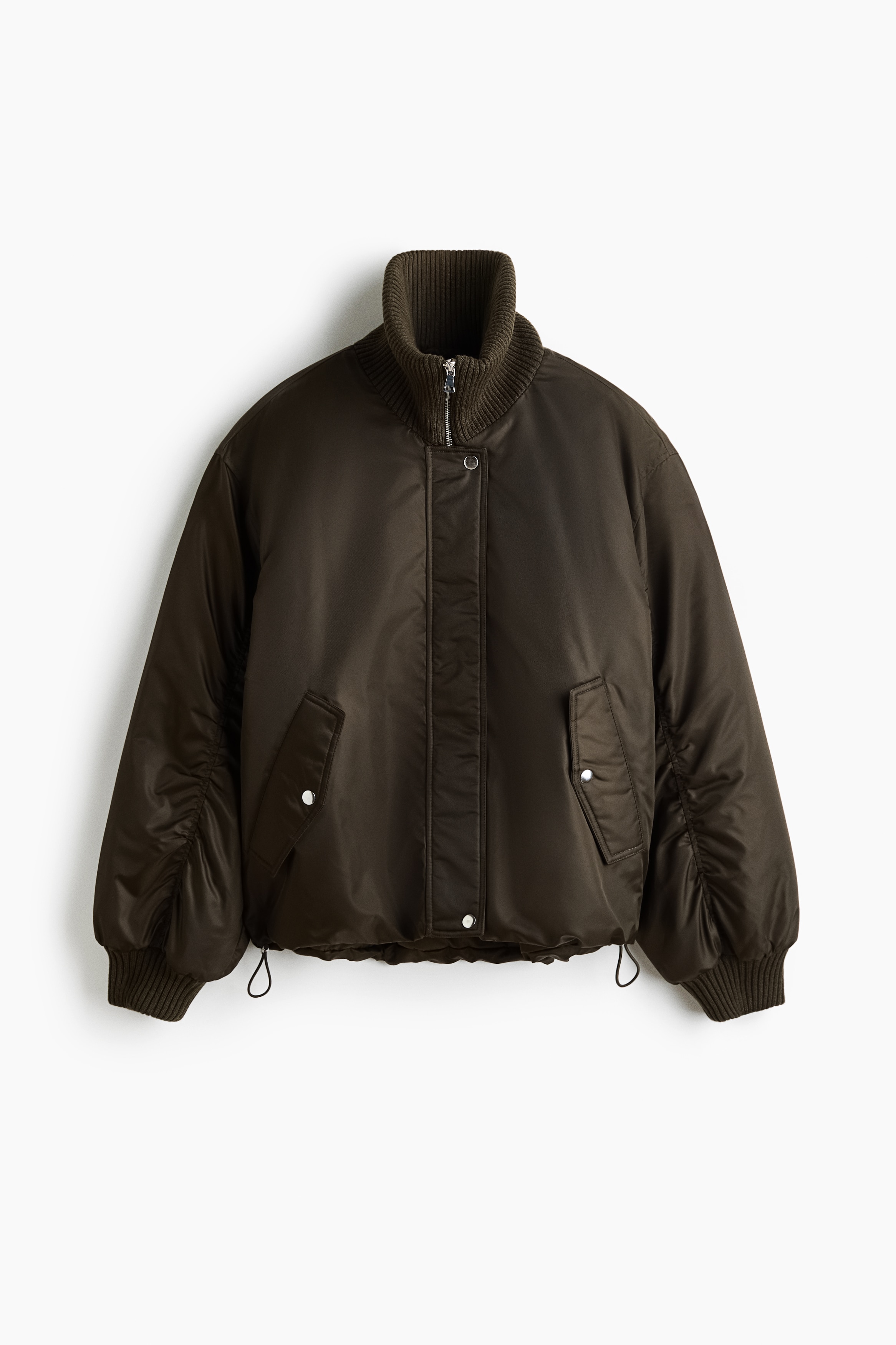 Ein oversized, dunkelbrauner, gesteppter Bomberjacke wird vom Model getragen, wobei die HÃ¤nde in den seitlichen Pattentaschen stecken. Die Jacke verfÃ¼gt Ã¼ber einen hohen Rippkragen, Ã¼berschnittene Schultern und einen gerafften Saum und ist Ã¼ber einem cremefarbenen Rollkragenoberteil und einer schwarzen Wide-Leg-Hose gestylt.