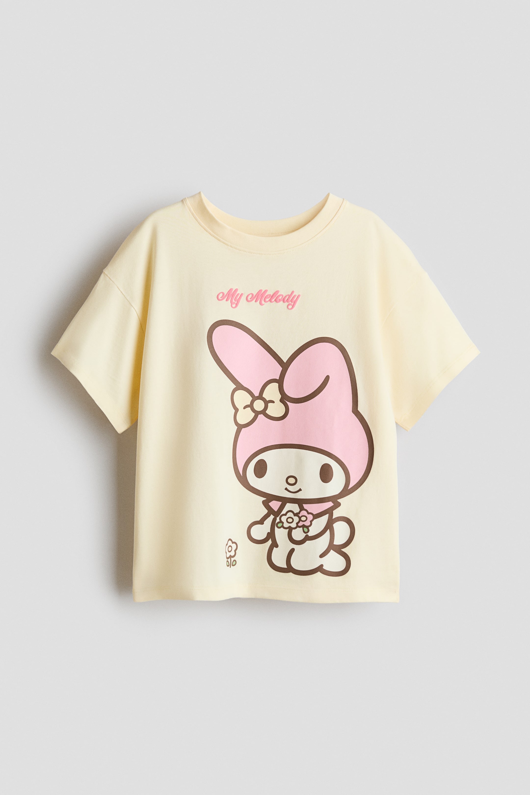 View larger image: The oversized t-shirt is shown from the front, featuring a light yellow soft cotton jersey with a round, ribbed neckline and dropped shoulders. It highlights a printed graphic of My Melody, outlined in brown with a pink hood and holding flowers, alongside pink "My Melody" text.