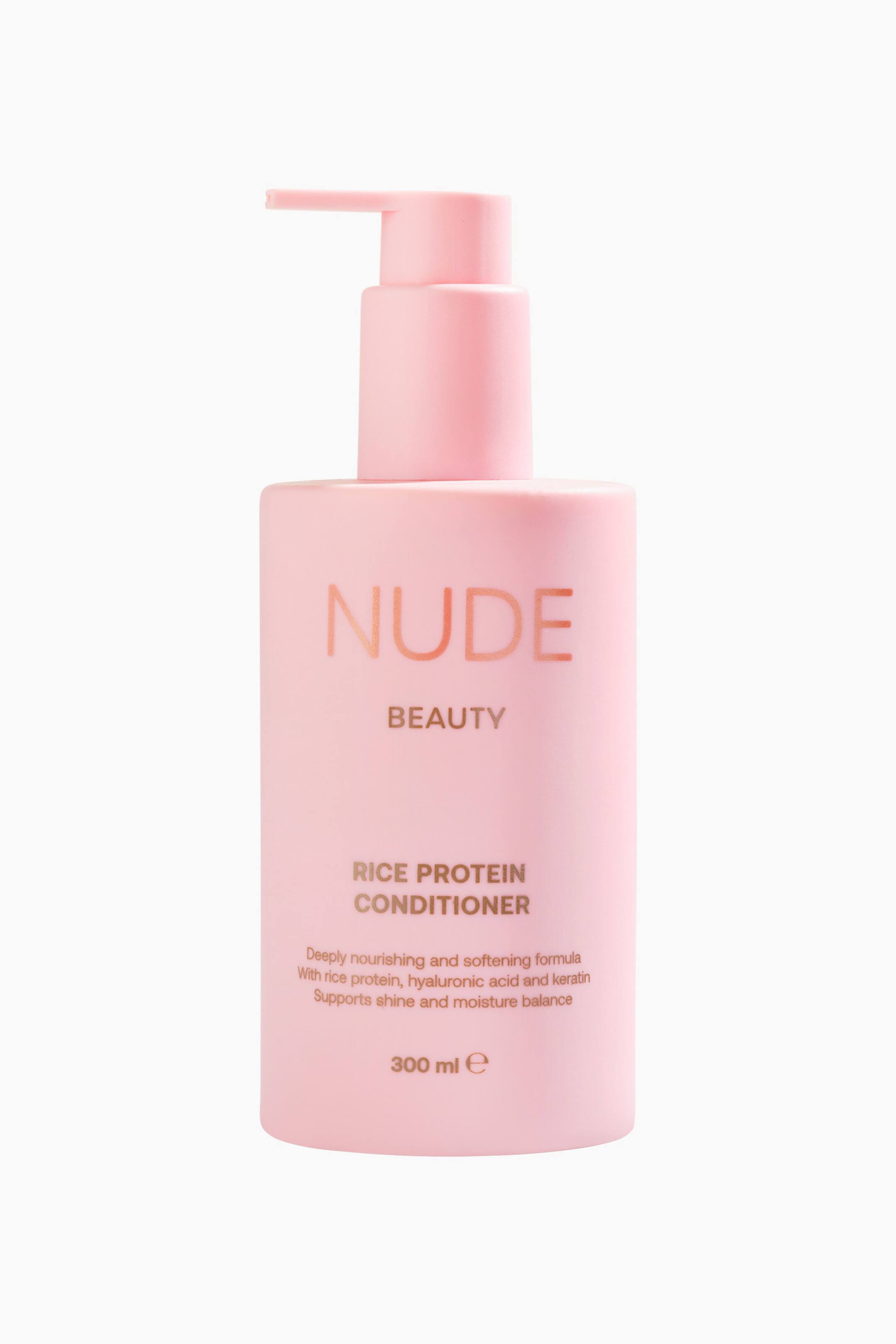 Vis større bilde: En rosa, rektangulær balsamflaske med avrundede kanter står frontvendt, med en matchende rosa pumpe-dispenser på toppen og viser 'NUDE BEAUTY RICE PROTEIN CONDITIONER' og '300 ml e' på forsiden.