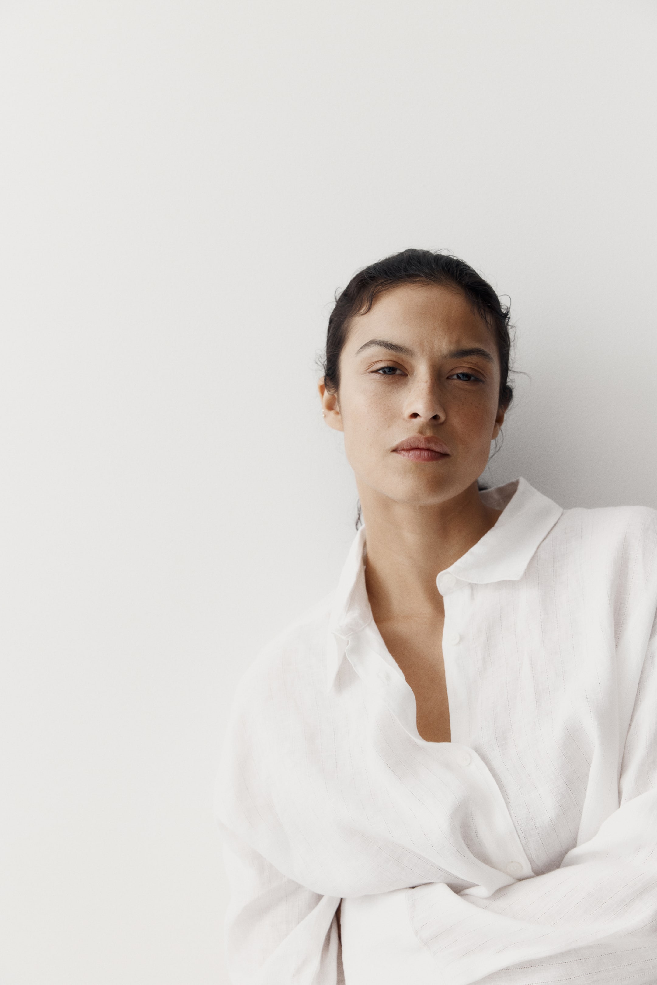 View larger image: A white collared shirt is worn, showcasing a subtle vertical textured weave and a pointed collar. The shirt's button-front placket is partially unbuttoned, revealing a white undershirt, and its long sleeves are visible with the arms crossed.