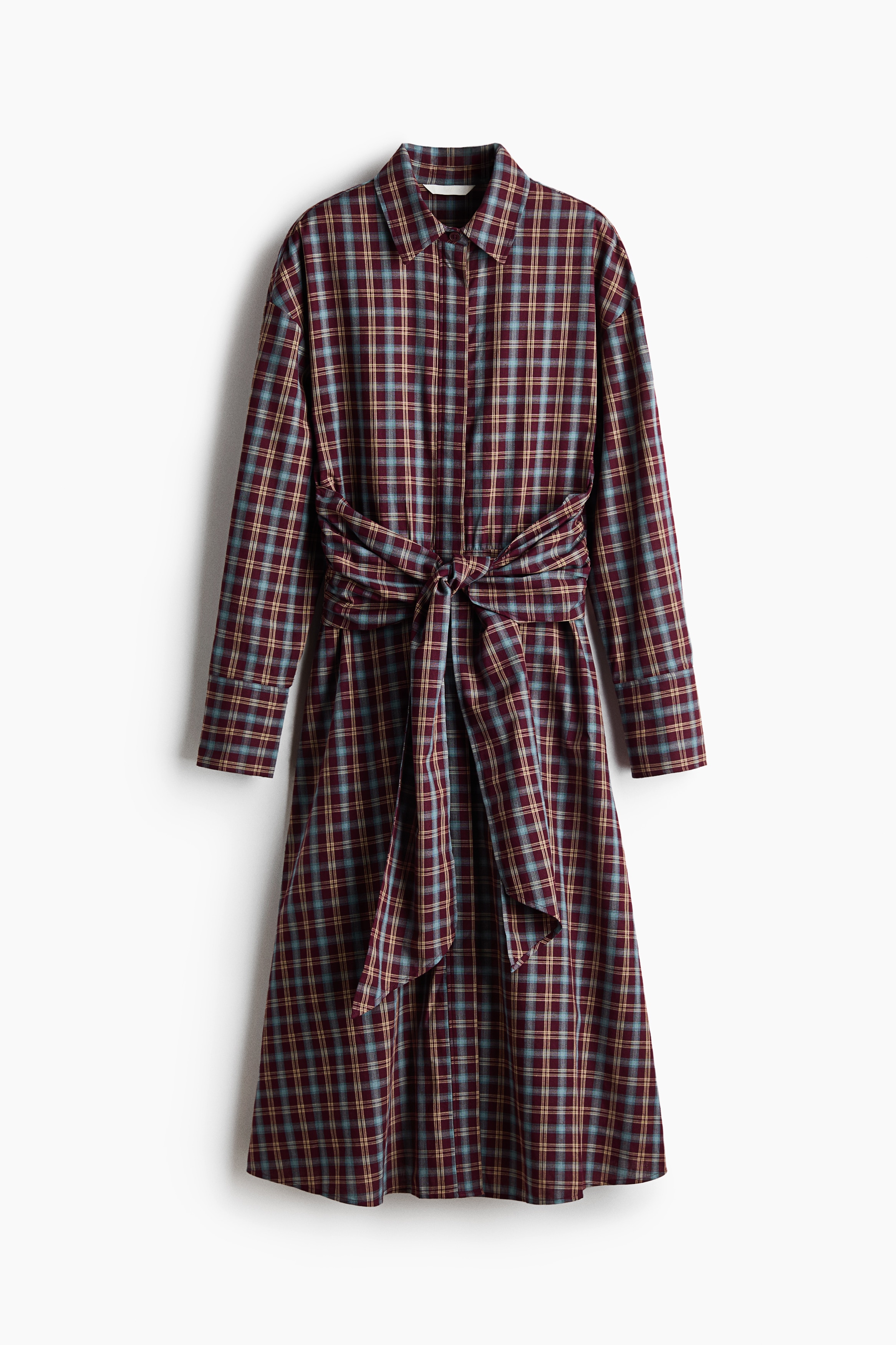 A plaid shirt dress in a burgundy, blue, and beige check pattern is being worn by the model. The dress features an open collar, dropped shoulders, long sleeves, and side panels tied in a knot at the front waist.