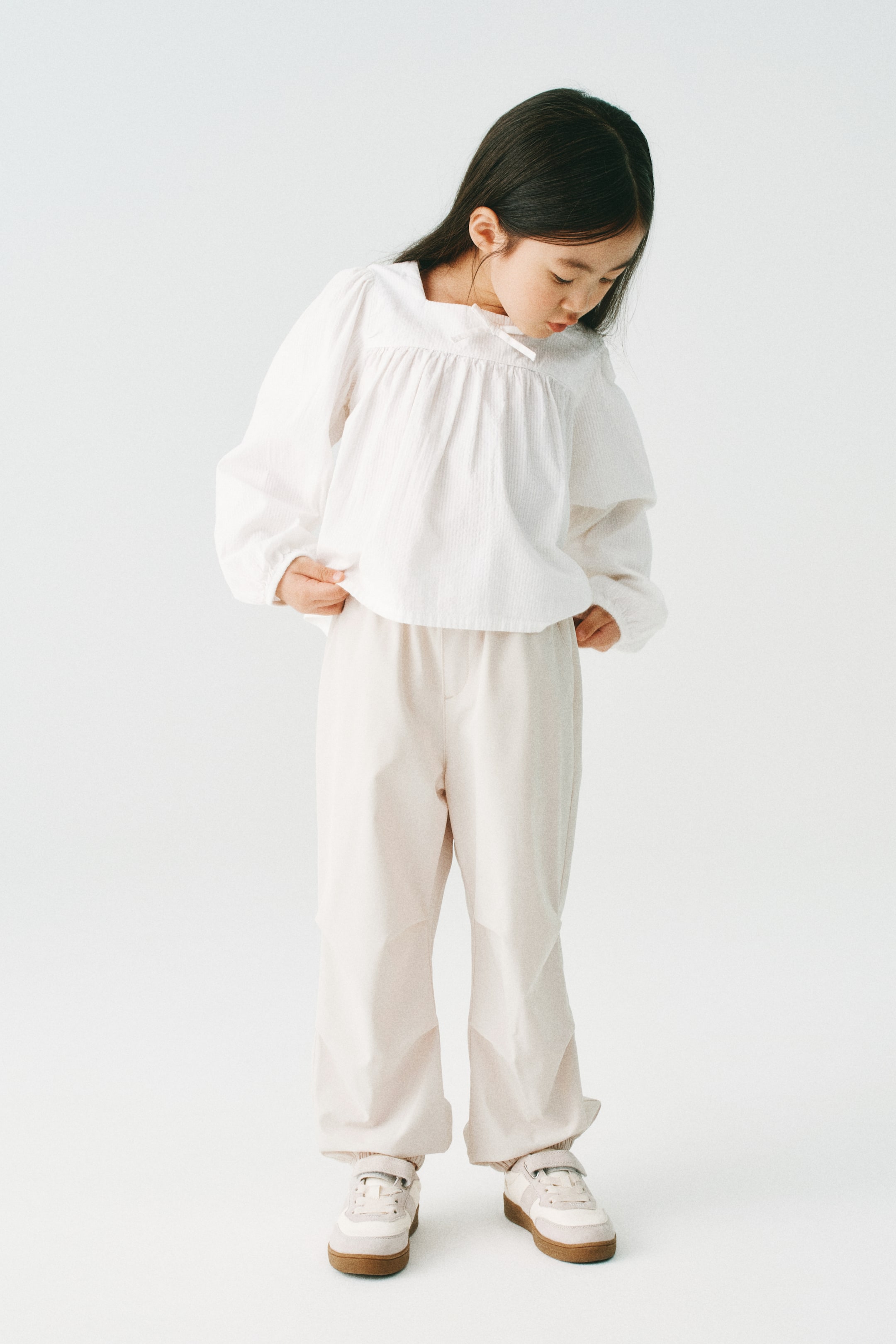 拡大画像を表示: 白い長袖ブラウスは、スクエアネックラインに小さなリボンが付いており、ボディス部分にはギャザーが施されています。袖はフルスリーブで、袖口にはゴムが入っています。このブラウスは、ウエスト部分にゴムが入ったライトベージュのゆったりとしたパンツと組み合わせられており、足首にはギャザーが施されています。